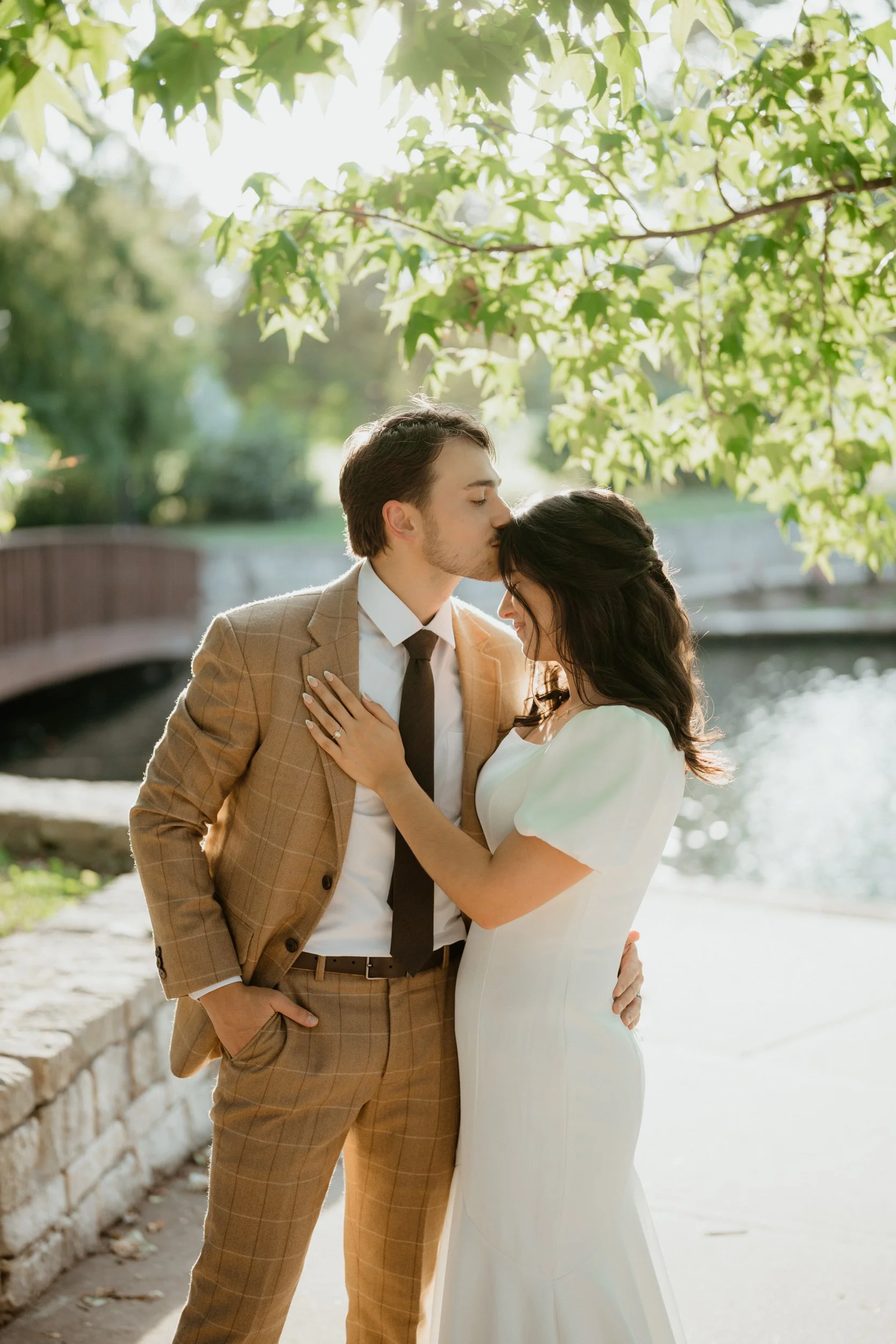A man in a brown plaid suit and a woman in a white dress sharing a tender moment outdoors by a river, with the man kissing the woman's forehead.