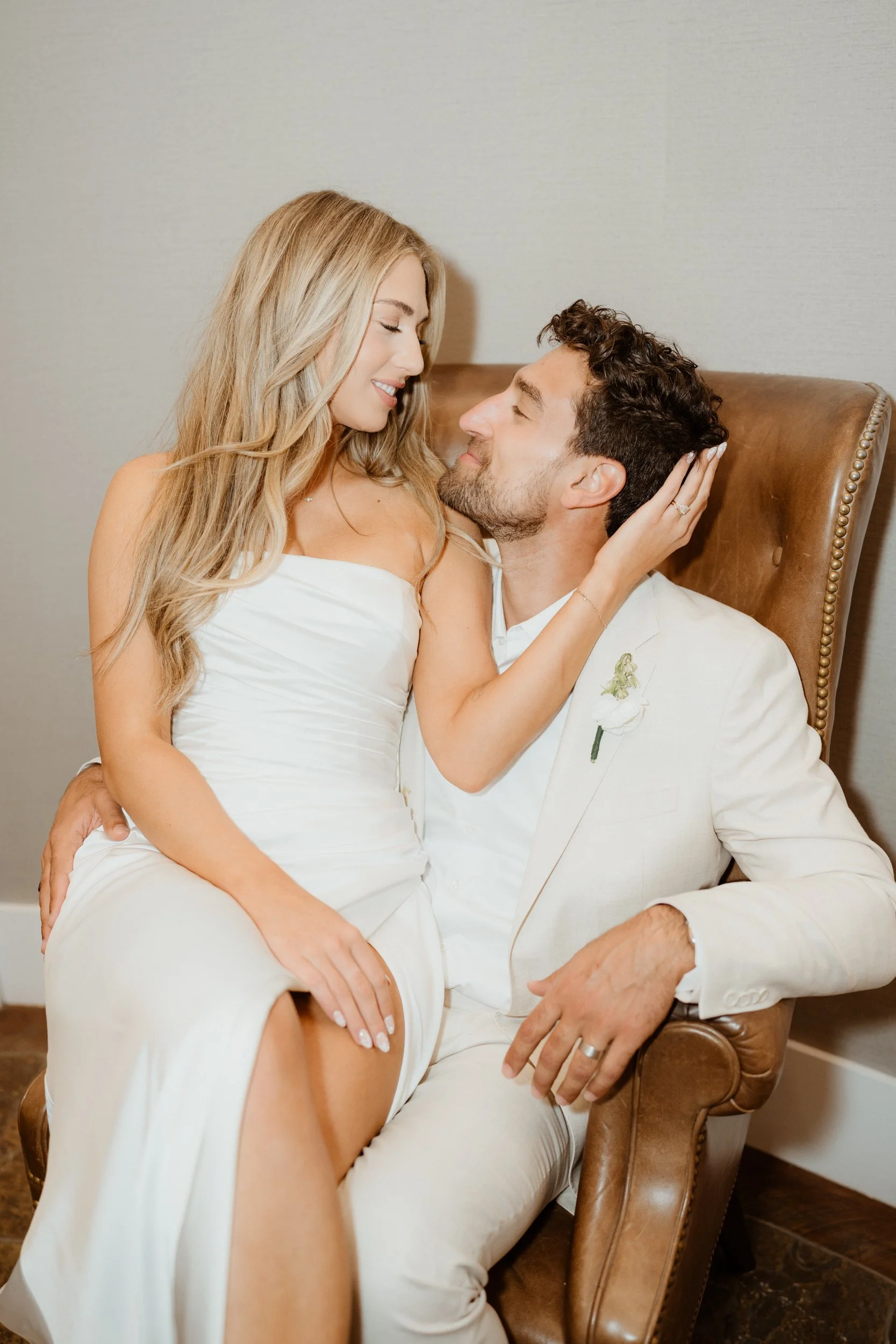 A woman in a white dress and a man in a white suit are sitting close together, smiling and looking into each other's eyes. The woman is touching the man's face while sitting on his lap in a cozy indoor setting.