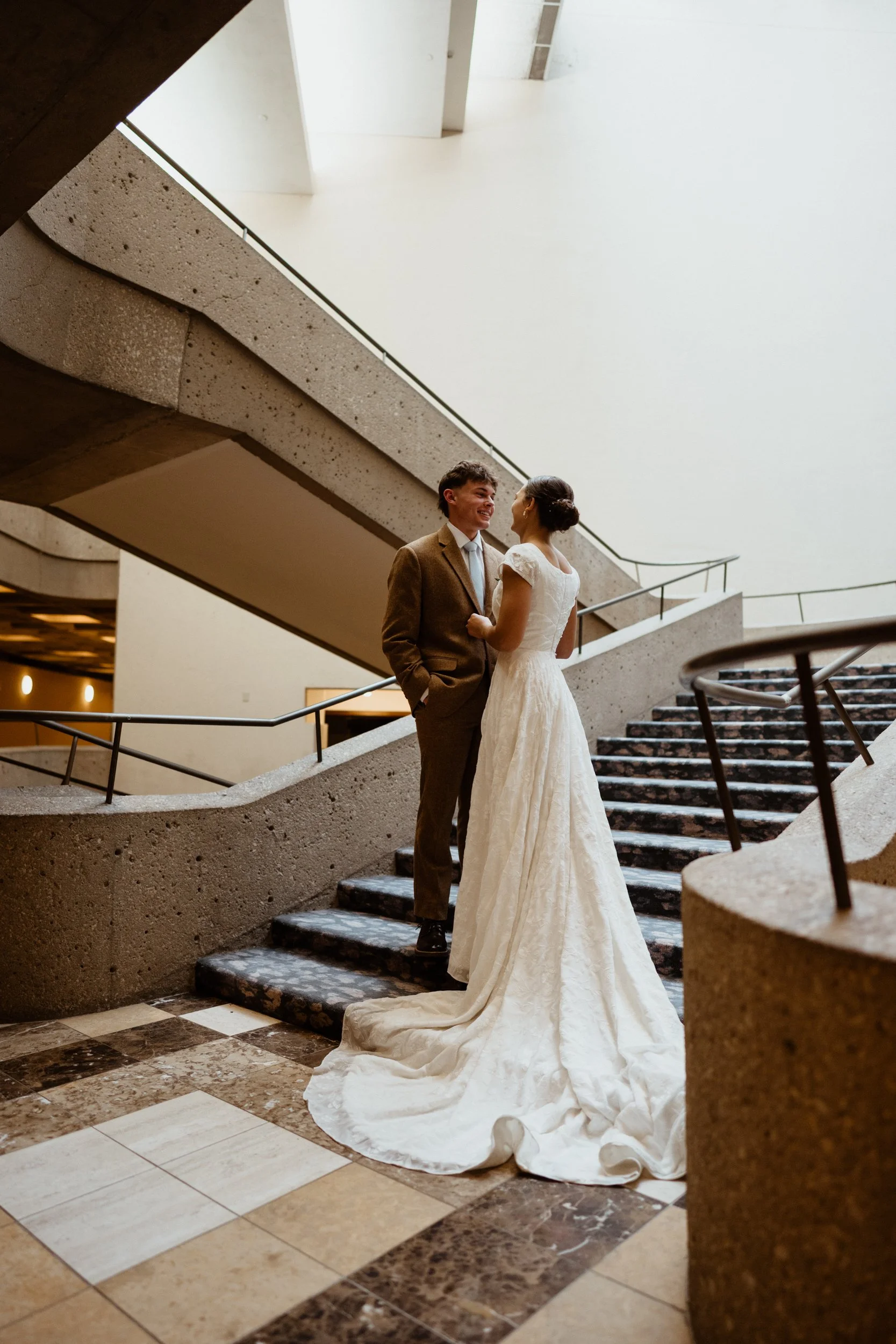 A wedding couple standing on a staircase in an indoor setting, with the bride in a long white gown and the groom in a brown suit, looking at each other and smiling.