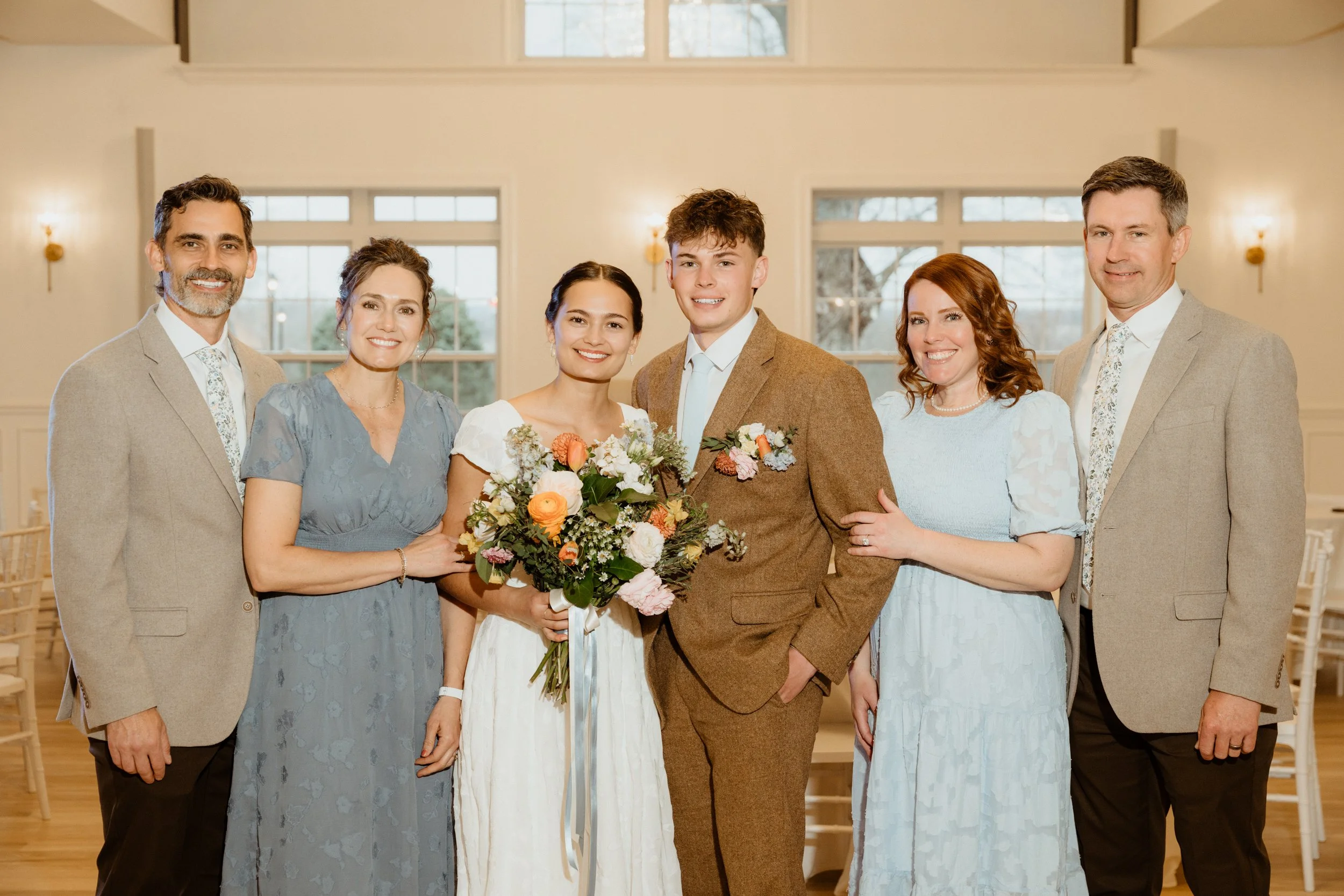 Group of seven people, including a bride and groom, standing inside a bright, elegant room with large windows. The bride holds a bouquet of flowers, and everyone is smiling.