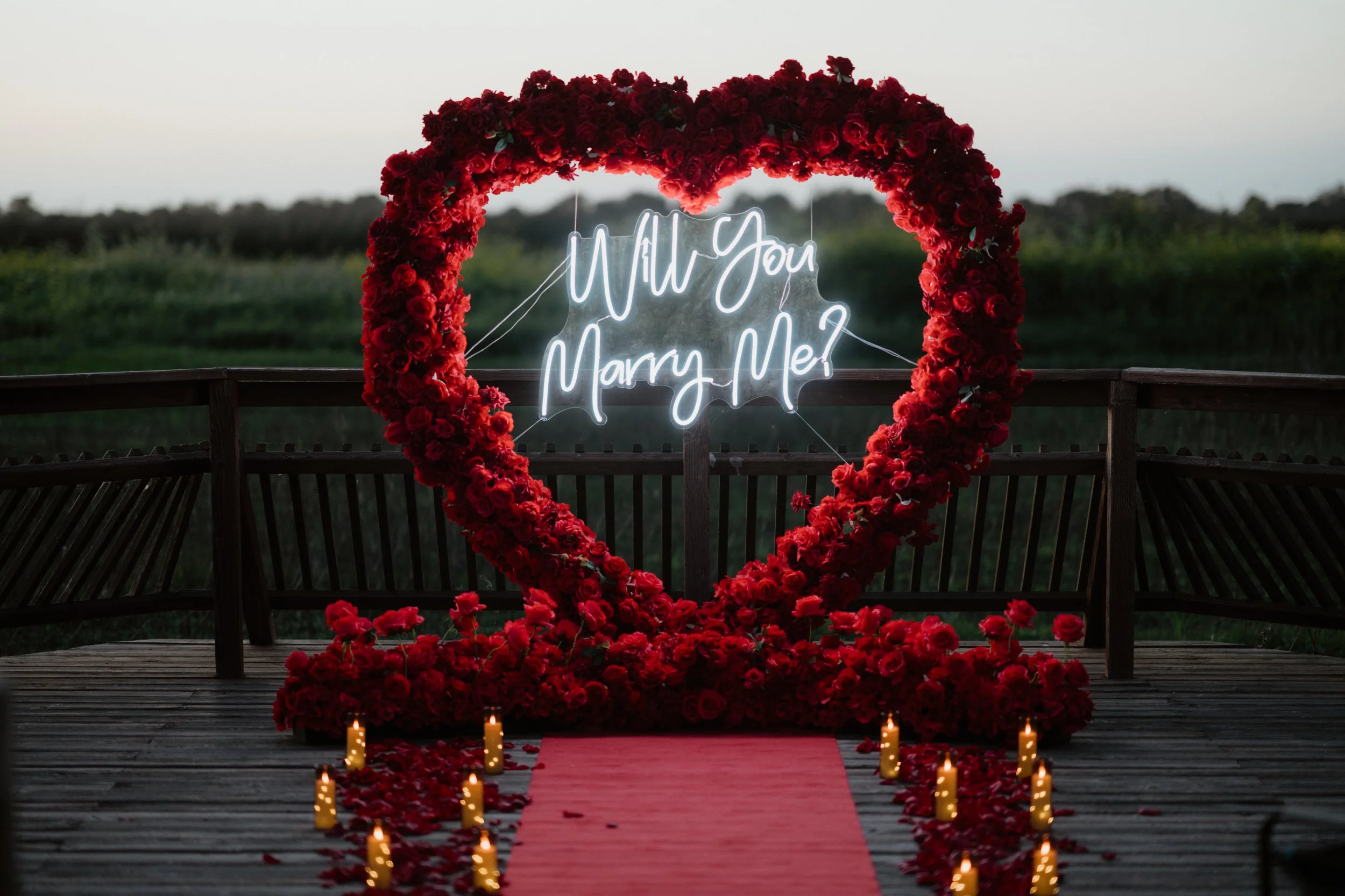 A large heart-shaped floral arch made of red roses on a wooden deck. Inside the arch, there is a neon sign reading 'Will You Marry Me?' in cursive. Small candles are placed along a red carpet leading up to the arch, with rose petals scattered on the carpet. The background shows a blurred natural landscape during dusk.