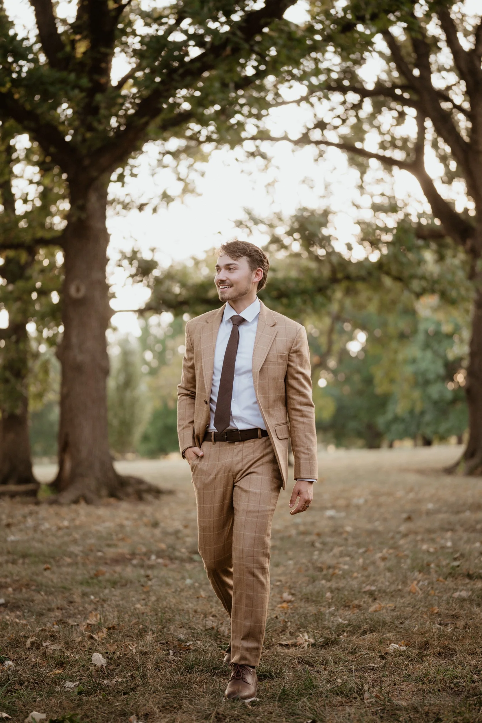 A man in a tan plaid suit walking outdoors through a park with large trees, smiling, during daytime.