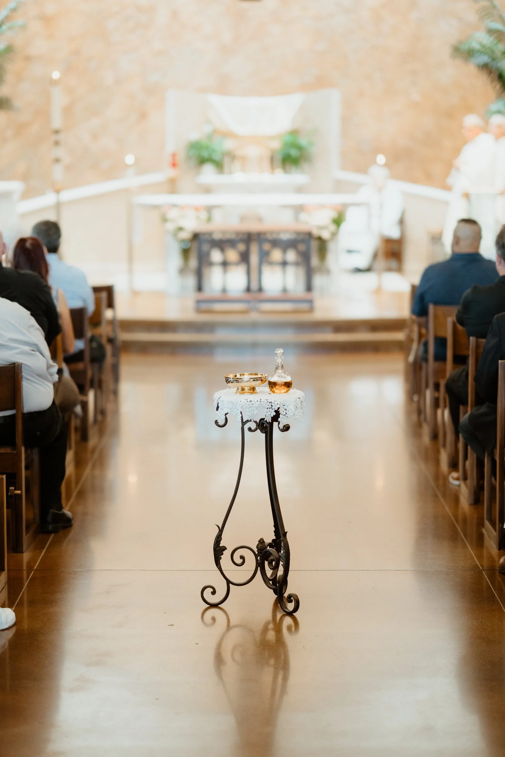 A small ornate table with a lace cloth, holding a decanter and bowl, in the aisle of a wedding venue with guests seated on both sides and an altar in the background.