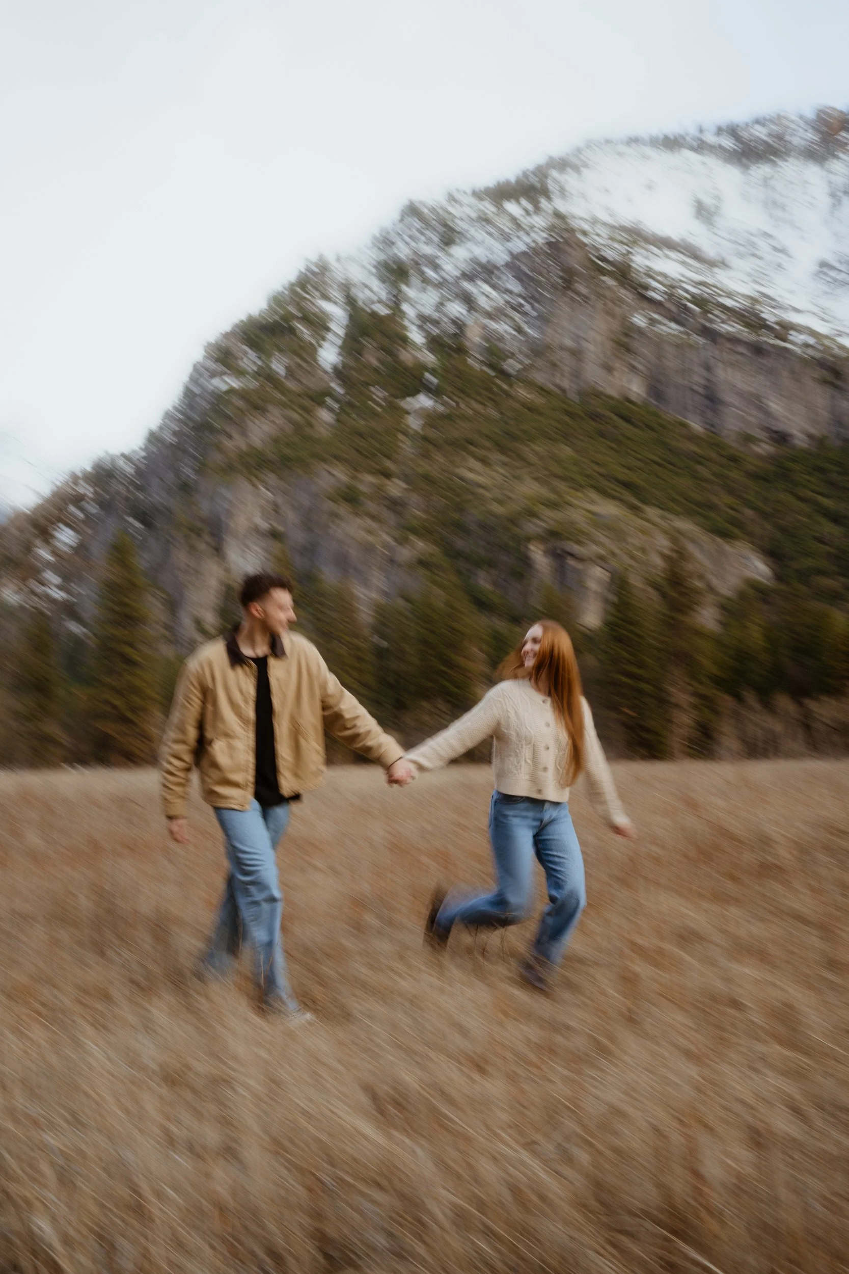 A couple holding hands and running through a field with a mountain in the background.