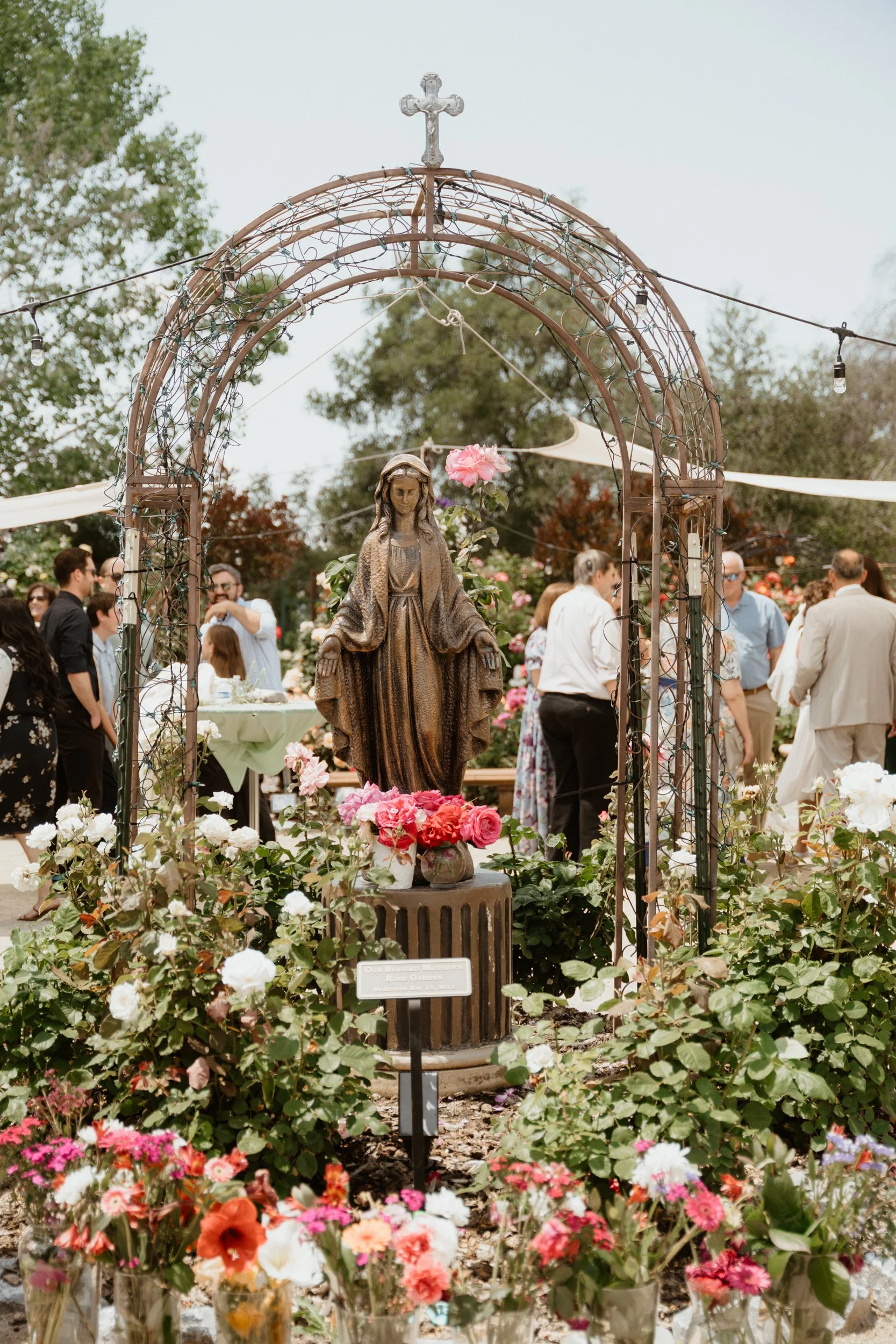 A statue of the Virgin Mary in a garden, surrounded by colorful flowers and a gathering of people in the background.