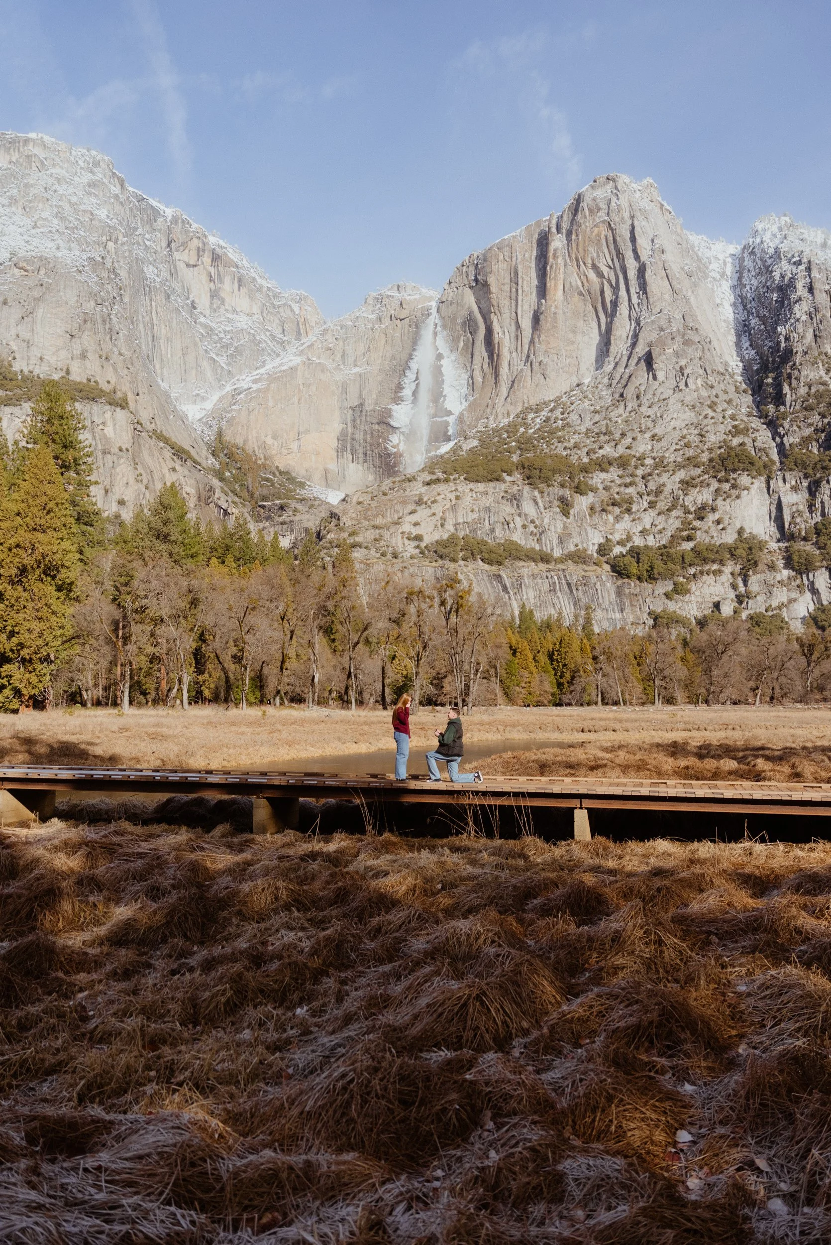 Two people on a wooden bridge with mountain scenery, trees, and a waterfall in the background.