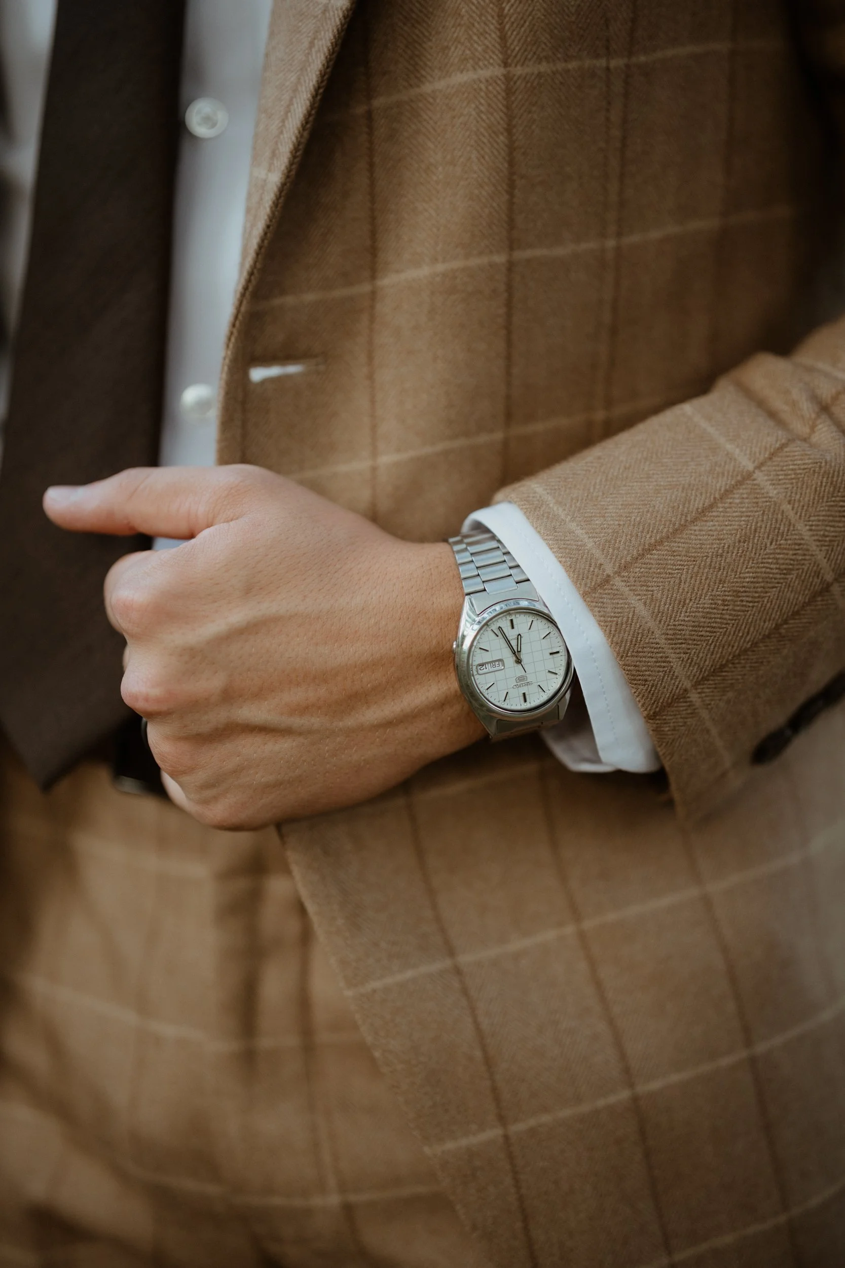 Close-up of a person's wrist wearing a silver watch with a white face, showing the time and day, with the person dressed in a brown checkered suit and white shirt.