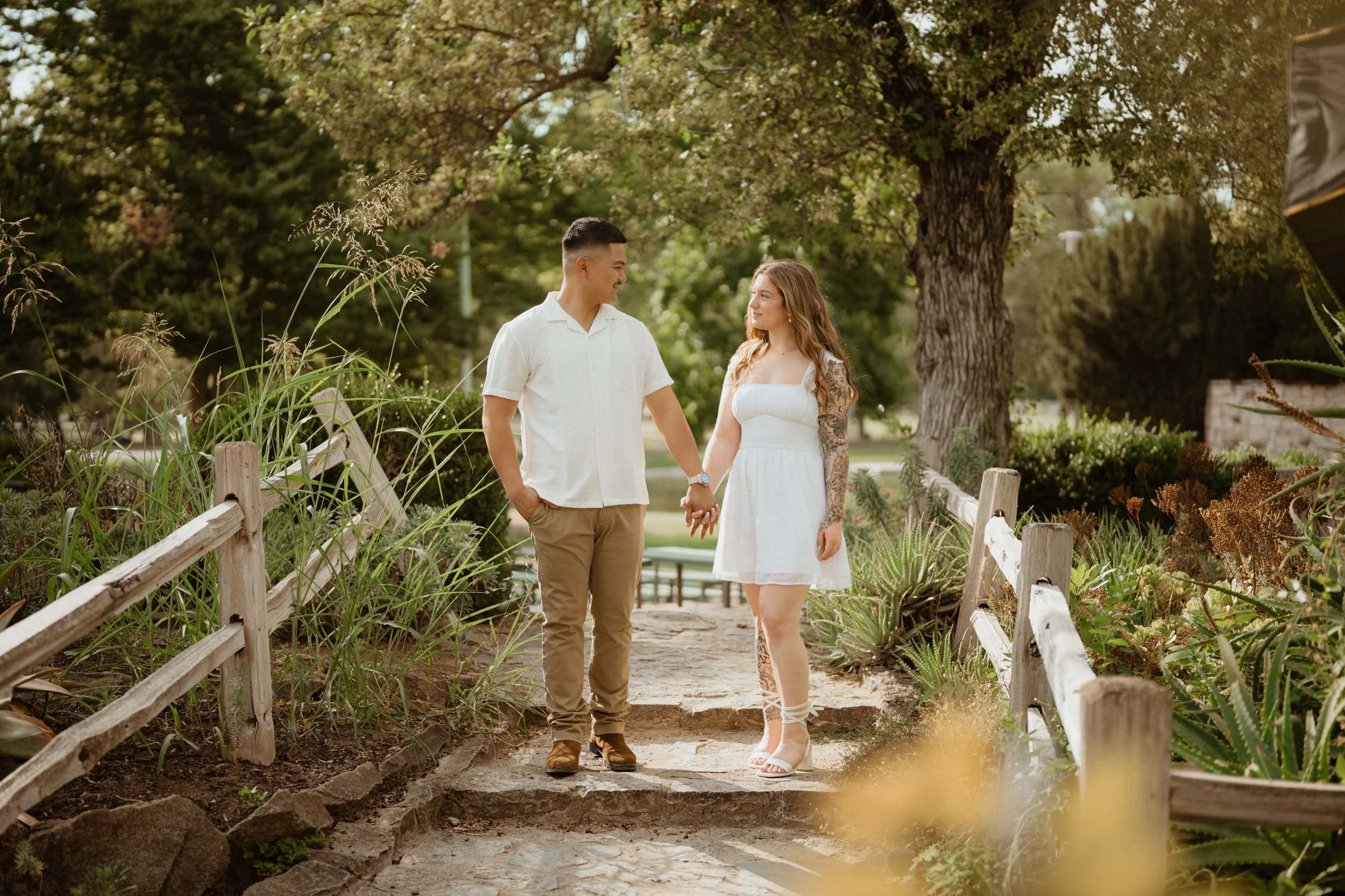 A couple holding hands and looking at each other on a stone pathway in a park with trees and plants.