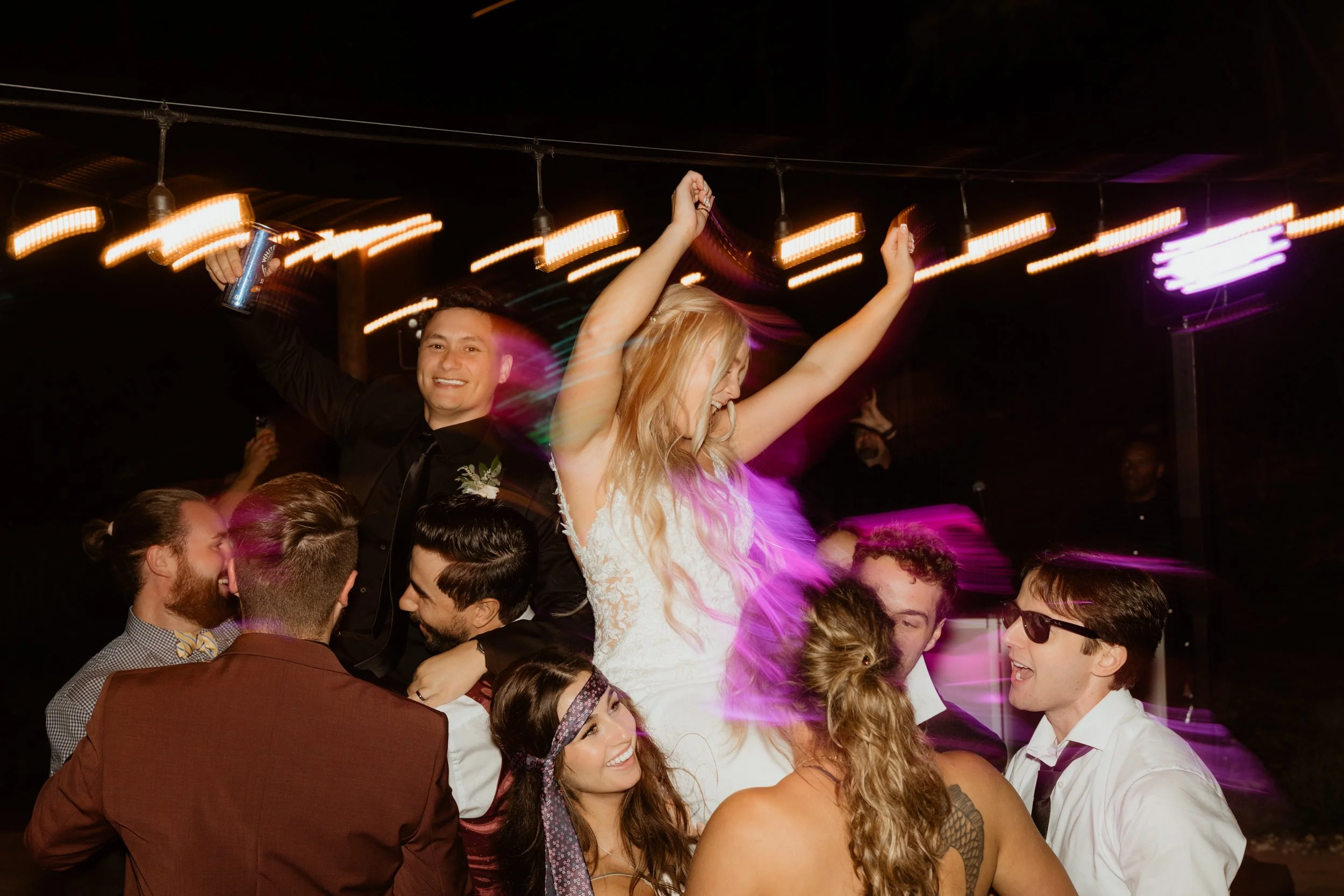 People celebrating at a wedding reception, with a bride and groom being lifted by friends, at night with string lights and colorful lighting effects.