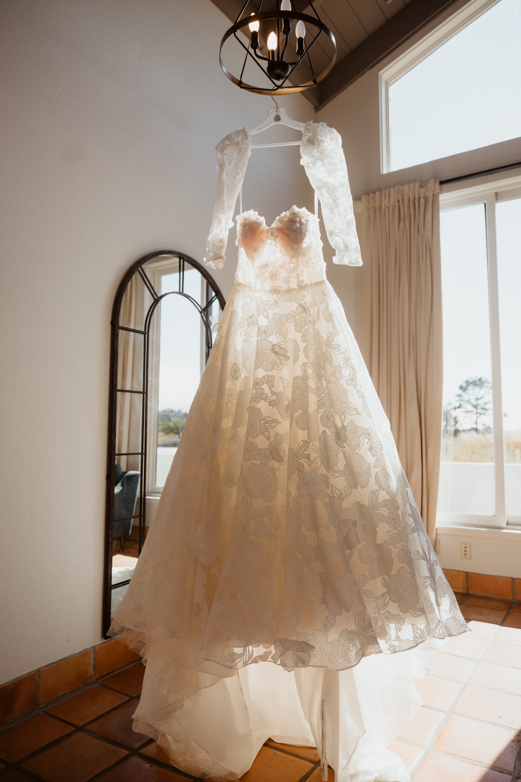 A wedding dress with lace overlay, hanging from a chandelier, displayed in front of a mirror near large windows in a room with tiled floor and cream-colored walls.