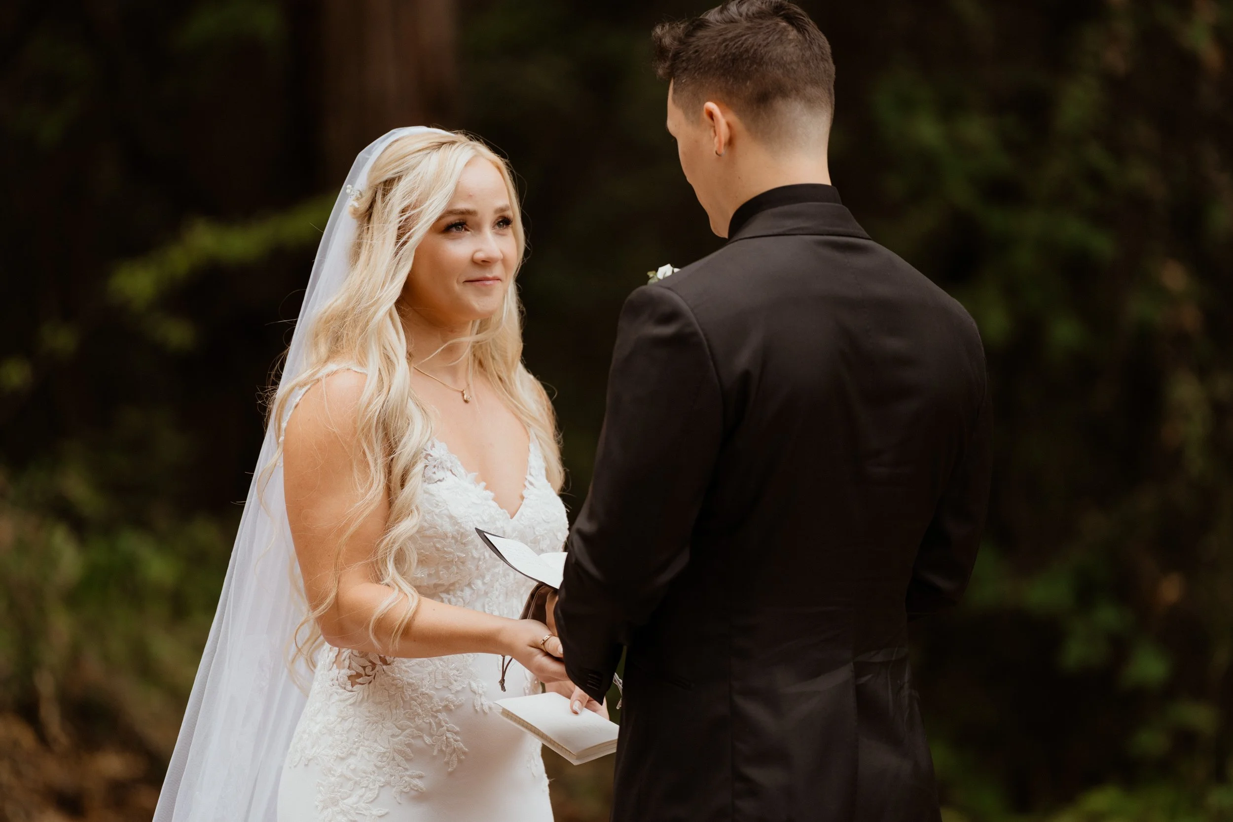 Bride and groom exchanging vows outdoors, with the bride holding a small book or note, surrounded by greenery.