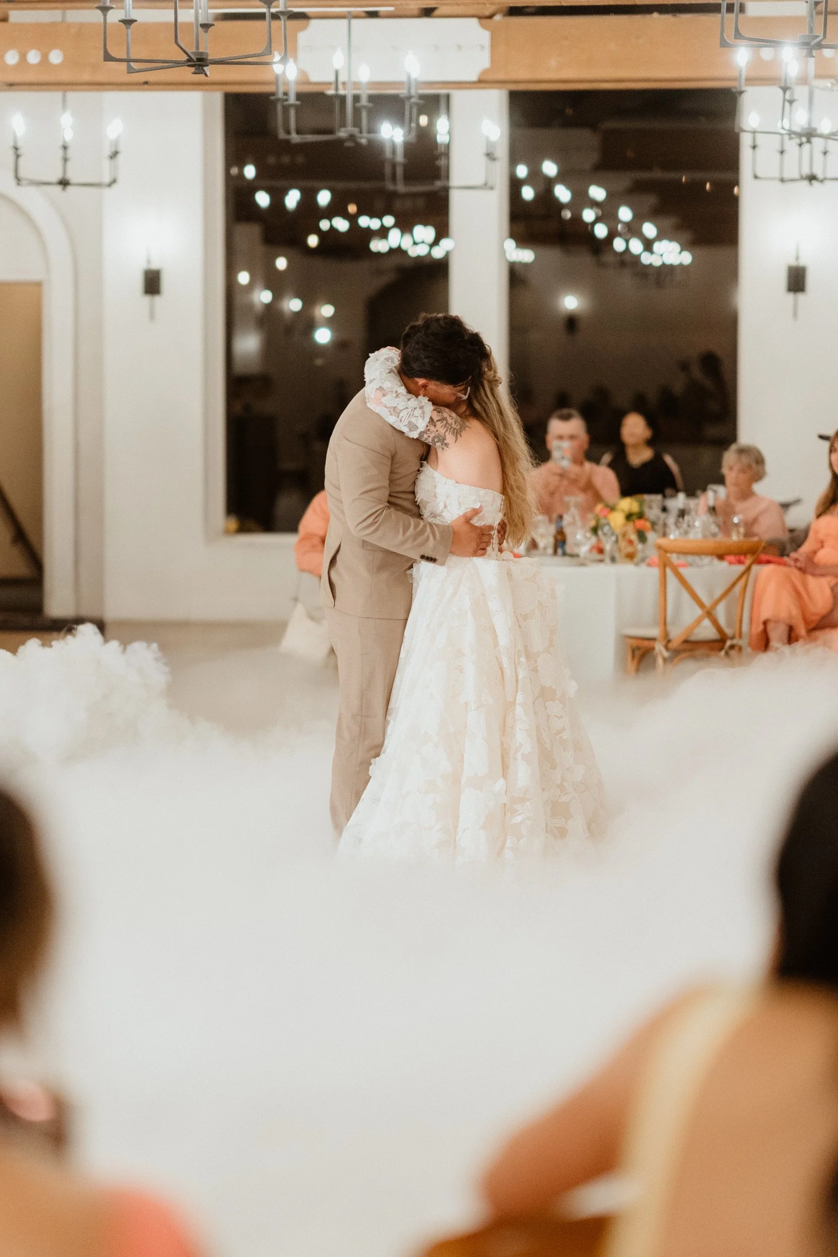 A couple shares a dance at their wedding reception, with guests seated around them and string lights overhead.
