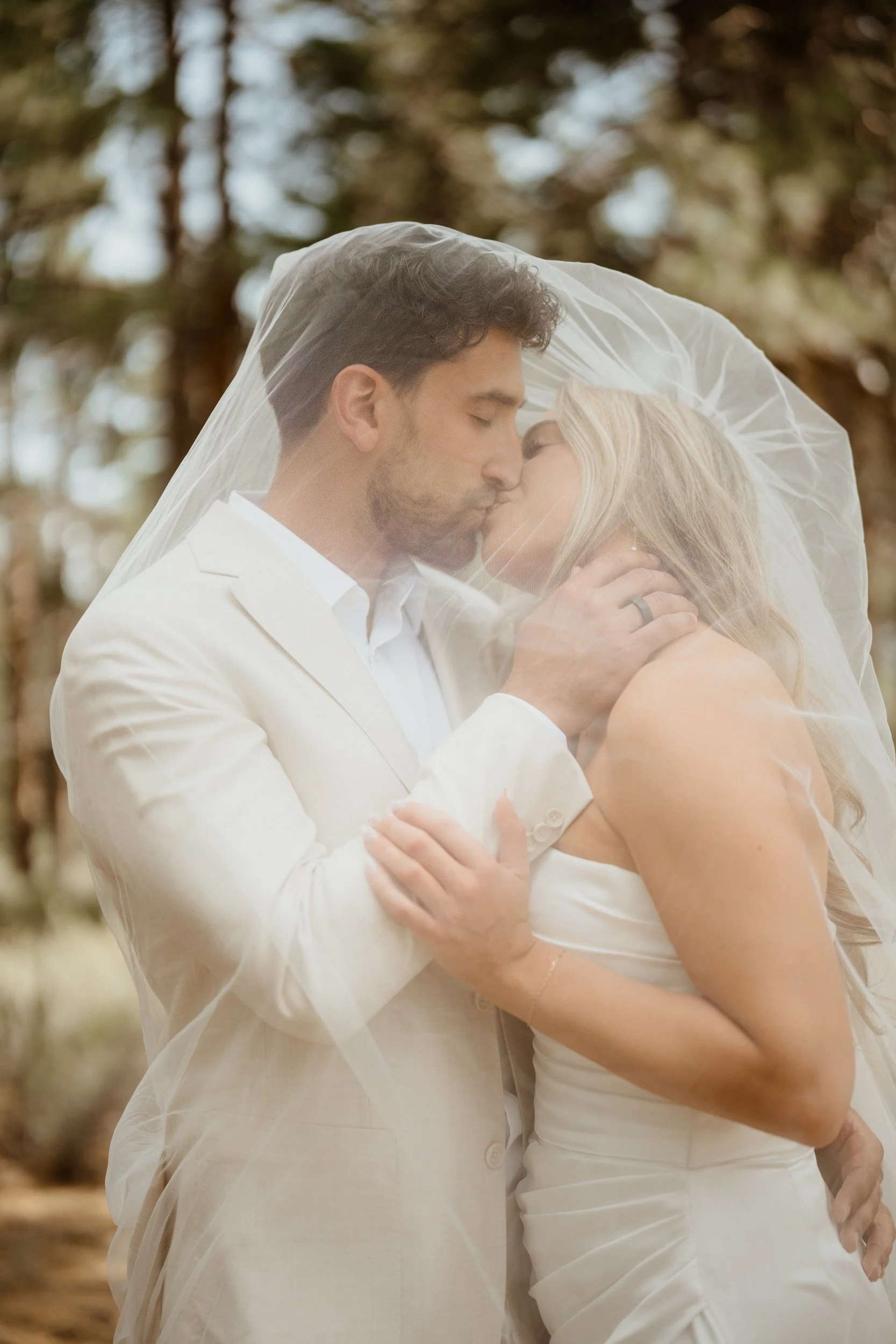 A newlywed couple sharing a kiss under a wedding veil in a forest setting.