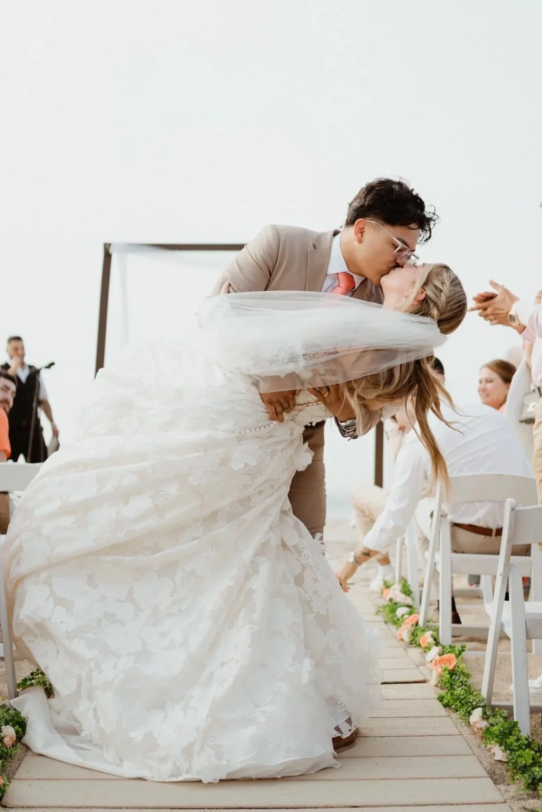 A couple, dressed in wedding attire, kiss during their outdoor wedding ceremony, with guests sitting nearby on a wooden aisle decorated with flowers.