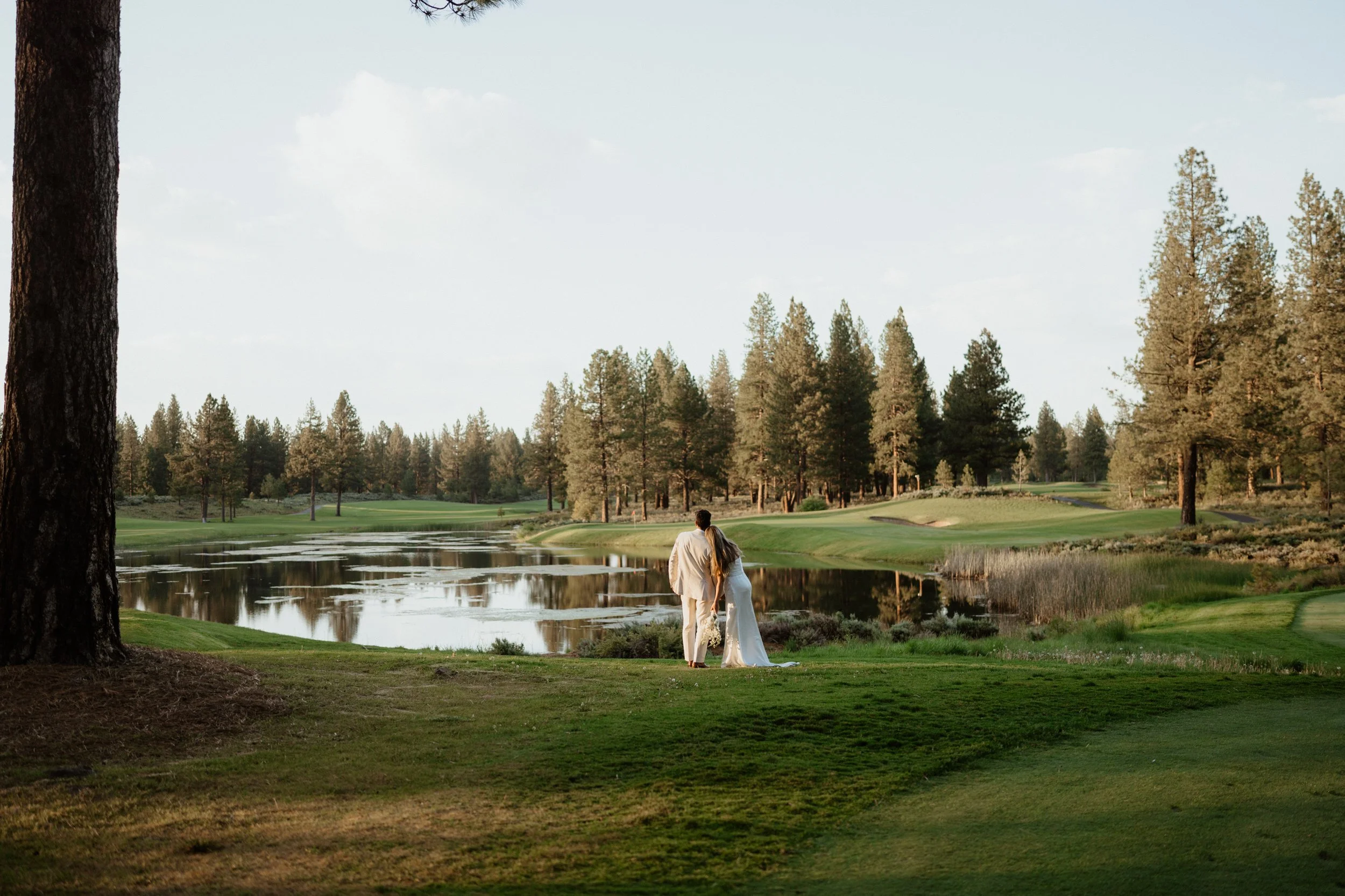 A couple dressed in wedding attire walking hand-in-hand along a grassy area by a lake on a golf course, with trees and a clear sky in the background.