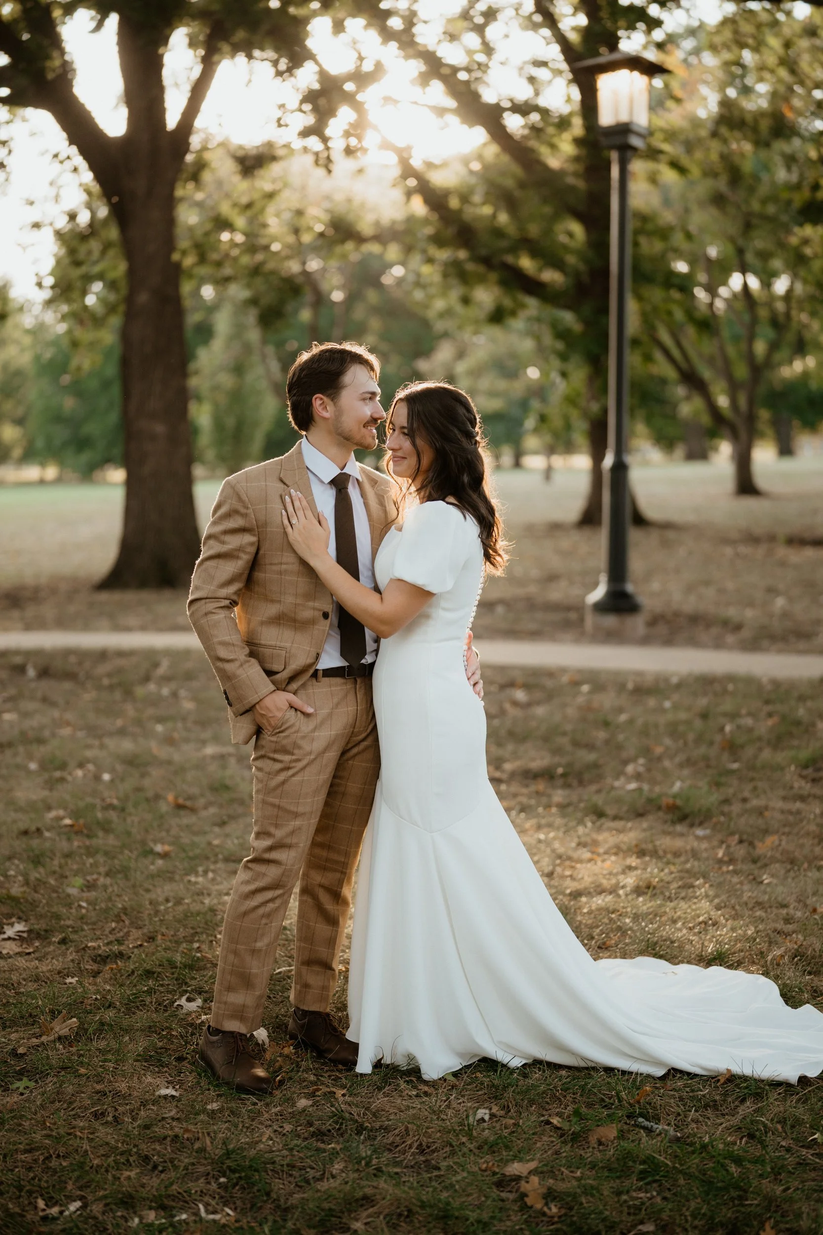 A couple dressed in wedding attire standing close together in a park with trees and a lamp post in the background, during sunset.