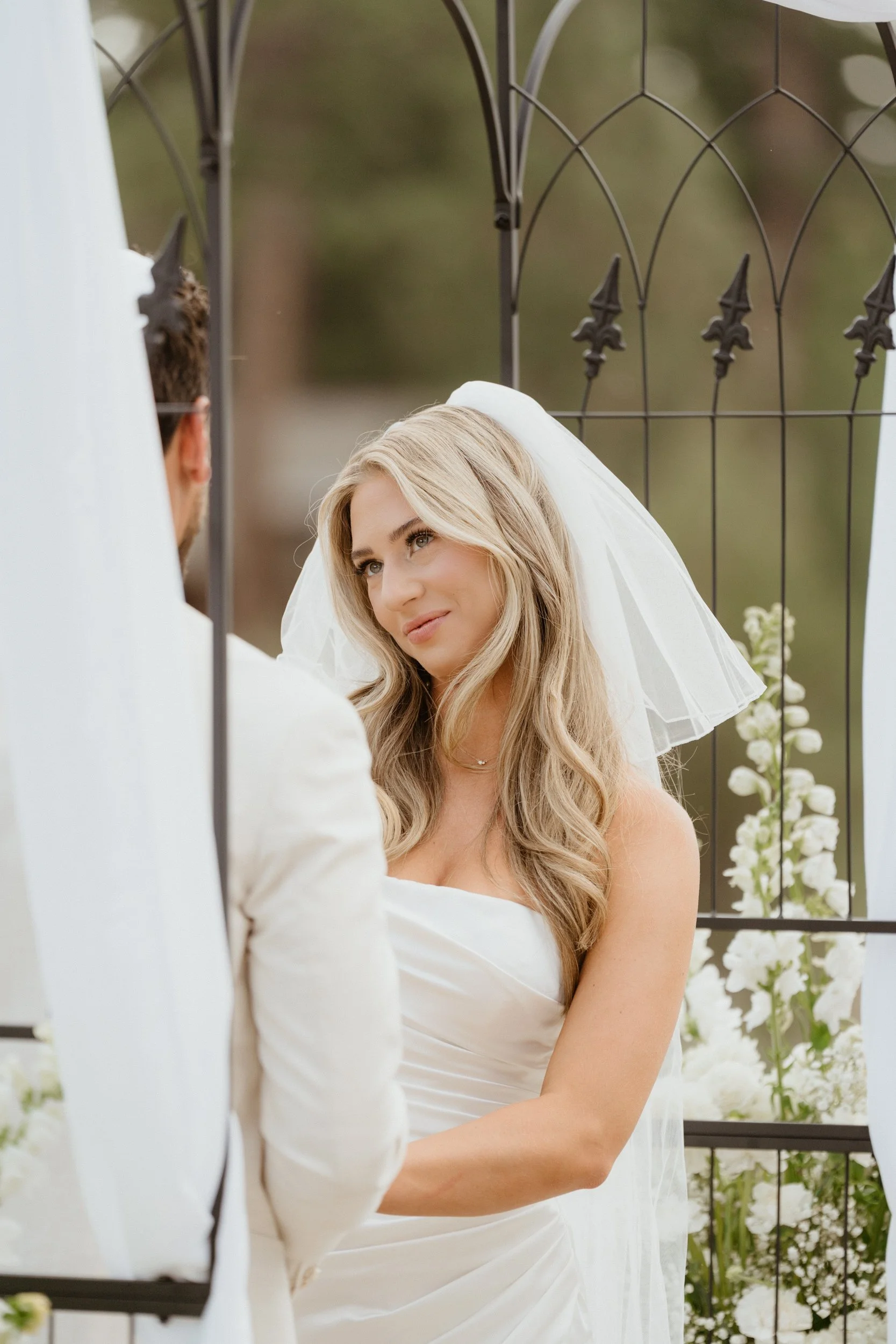 Bride and groom exchanging vows outdoors near a black metal gate decorated with white flowers, with the bride in a white wedding dress and veil, and the groom in a white suit.