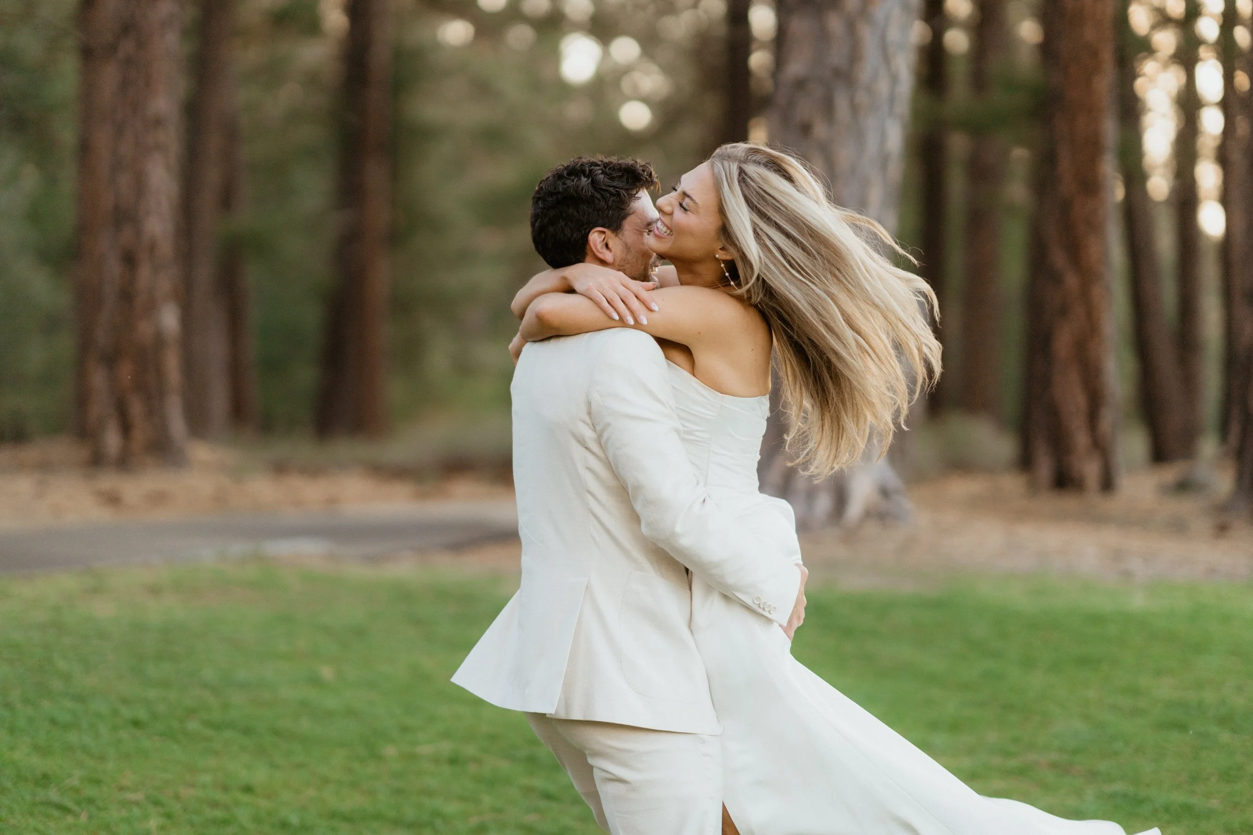 A joyful couple dressed in wedding attire embracing and smiling in a forested area with tall trees and green grass.
