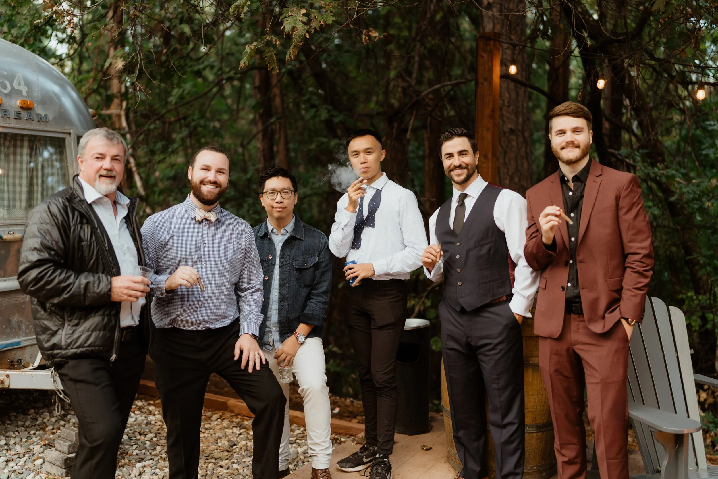 Group of seven men standing outdoors in a wooded area, dressed in semi-formal and casual attire, some holding drinks and a cigarette, with a vintage trailer and string lights in the background.