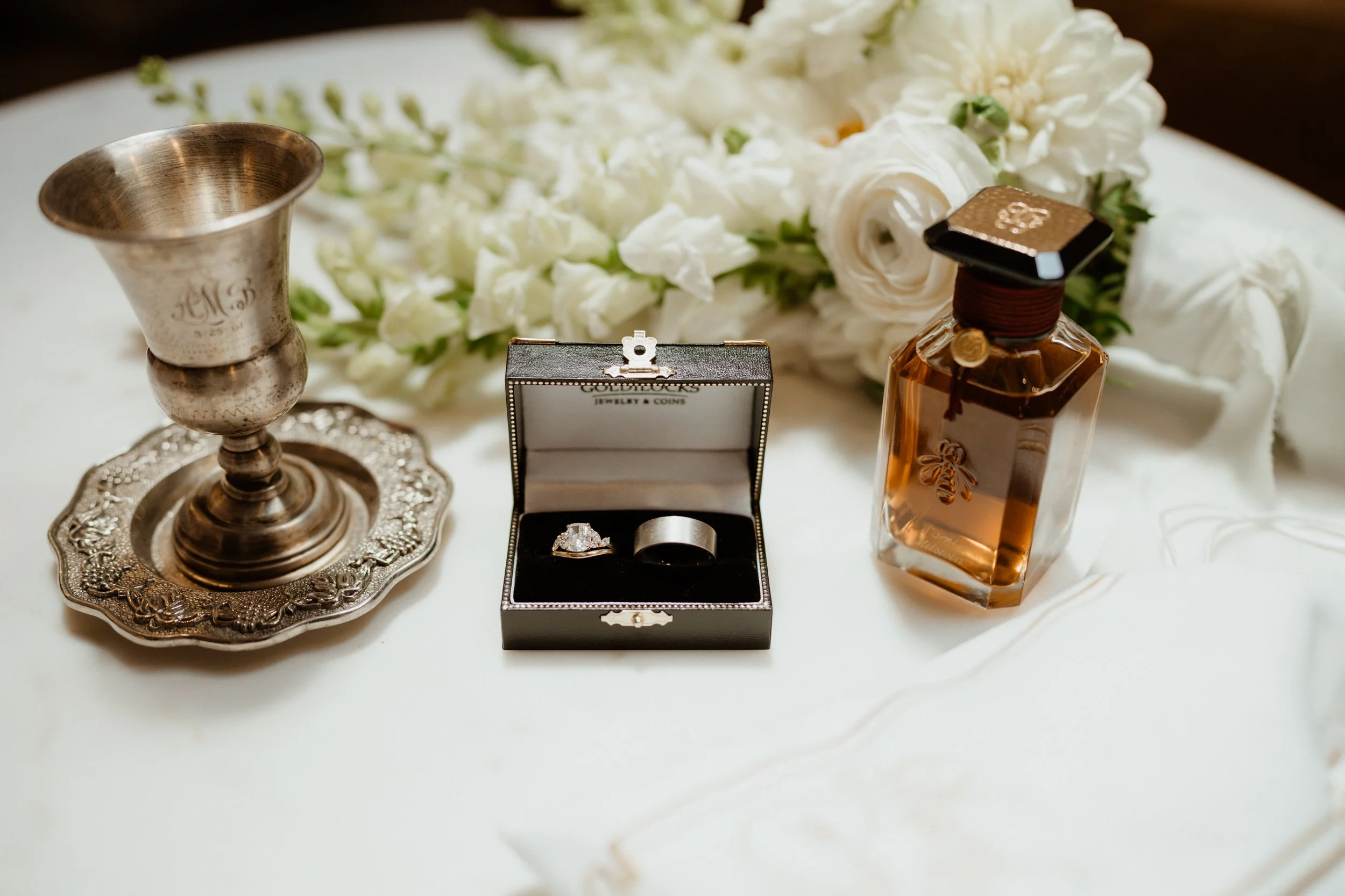 A collection of wedding items including a silver chalice on an ornate silver tray, a box with a large engagement ring and a wedding band, a bottle of perfume, and white flowers on a white cloth.