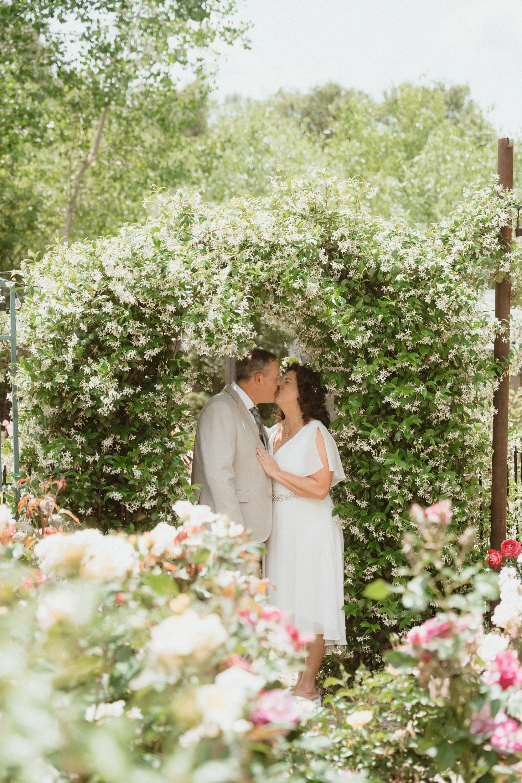 A couple in wedding attire sharing a kiss beneath a floral arch in a garden setting.