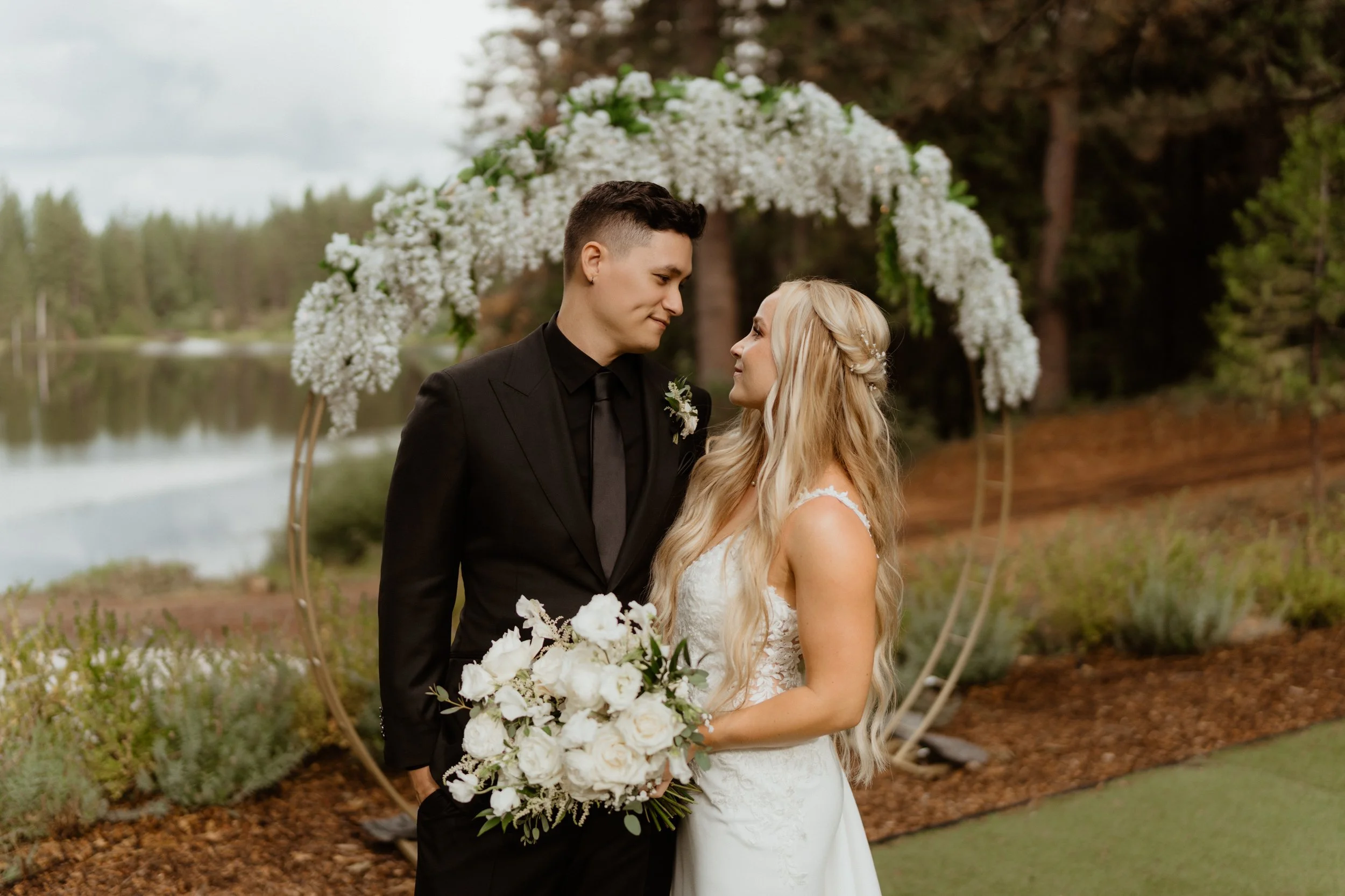 A bride and groom stand close together under a floral arch, looking into each other's eyes. The bride has long blonde hair and holds a bouquet of white flowers, wearing a white wedding dress. The groom wears a black suit and tie. The background features a lake surrounded by trees.