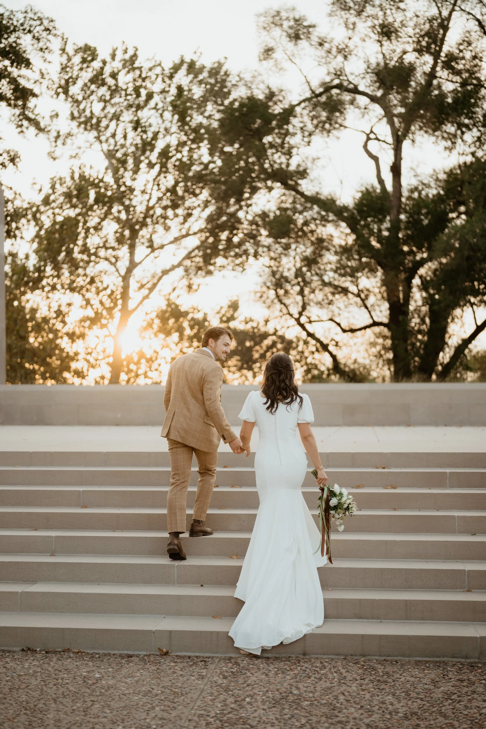 A bride and groom walking hand in hand up outdoor stairs during sunset, with trees in the background.