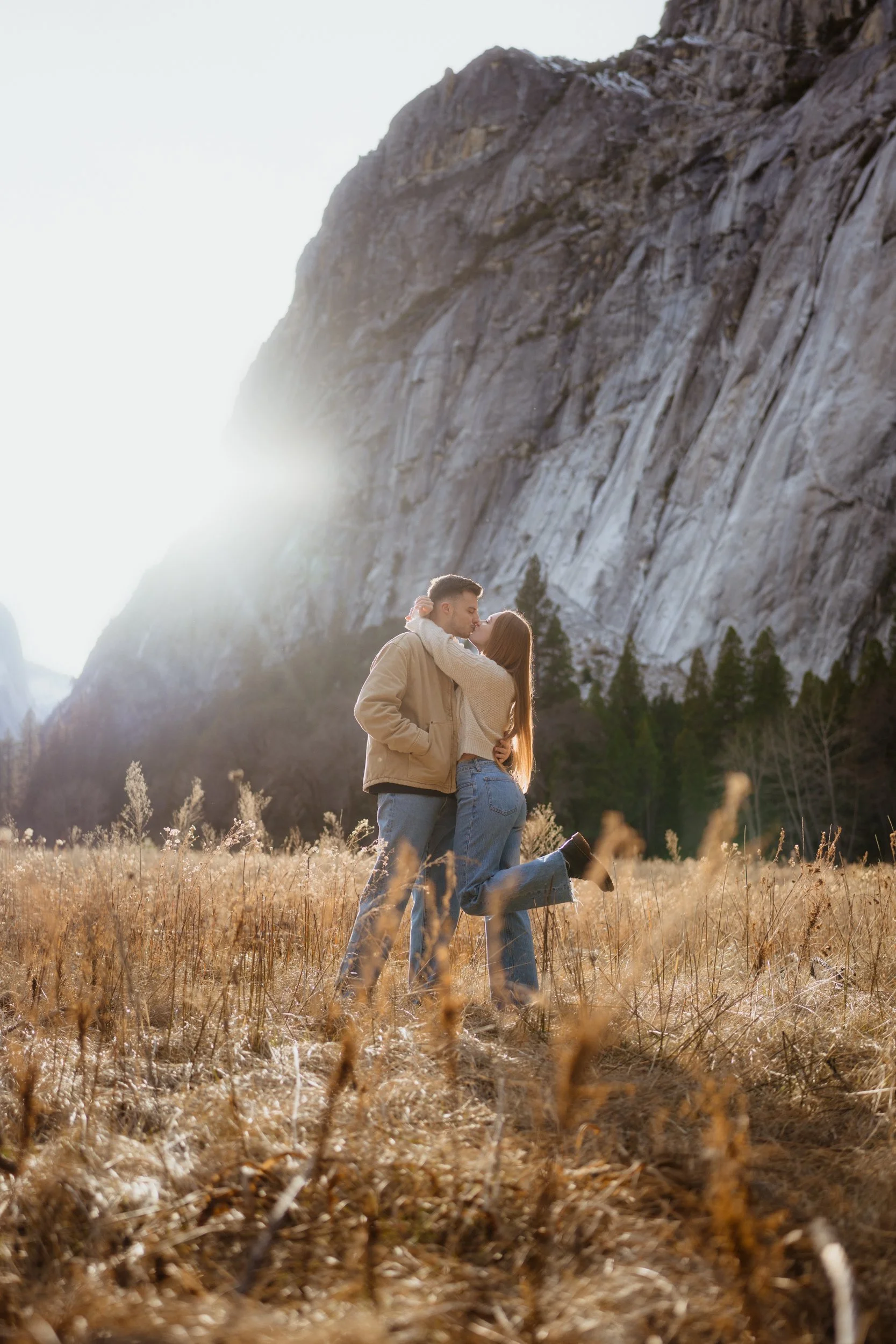 A couple embraces and kisses in a field of tall, dry grass with a mountain and trees in the background during sunset.