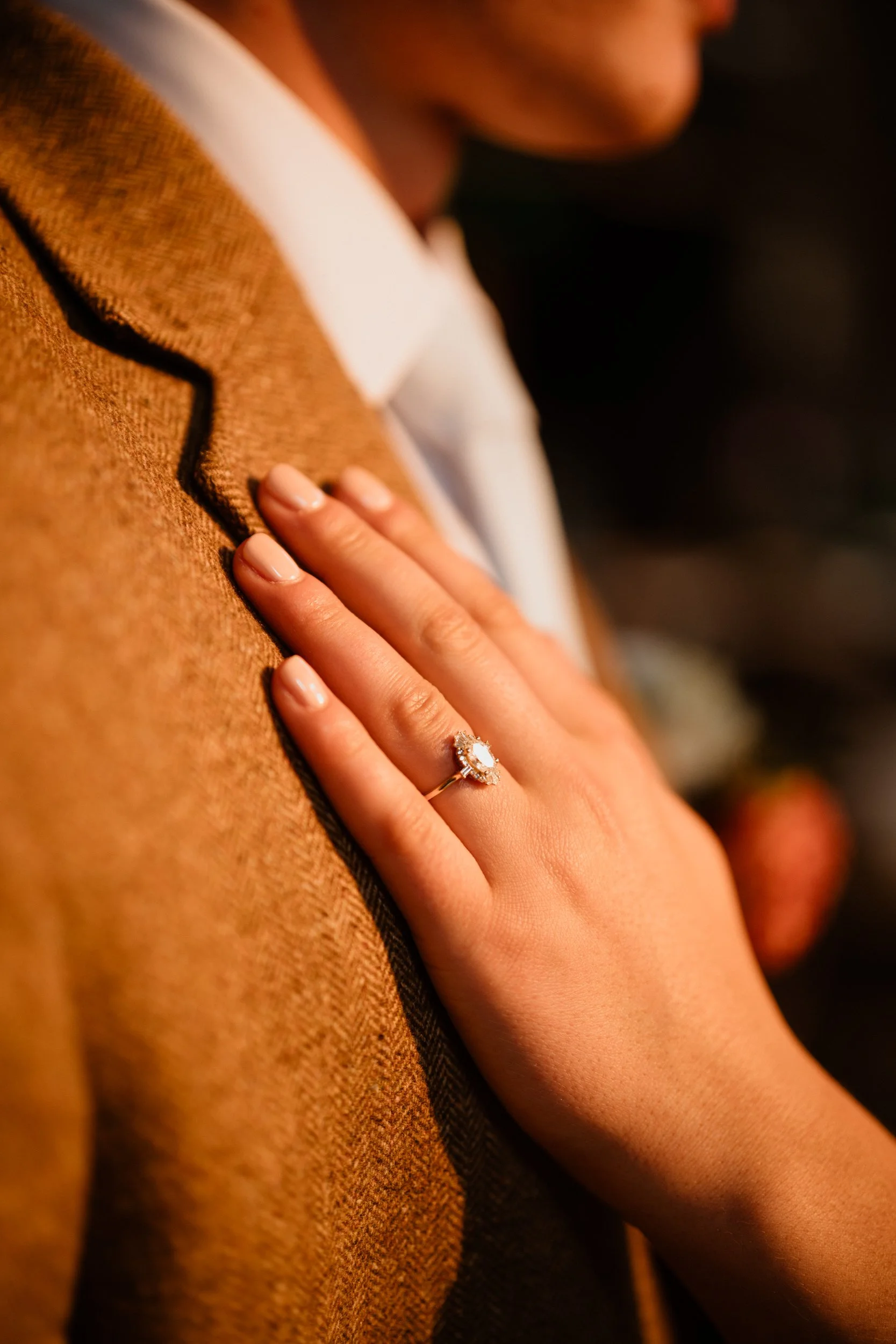 Close-up of a woman's hand with an engagement ring resting on a man's chest in a brown suit.