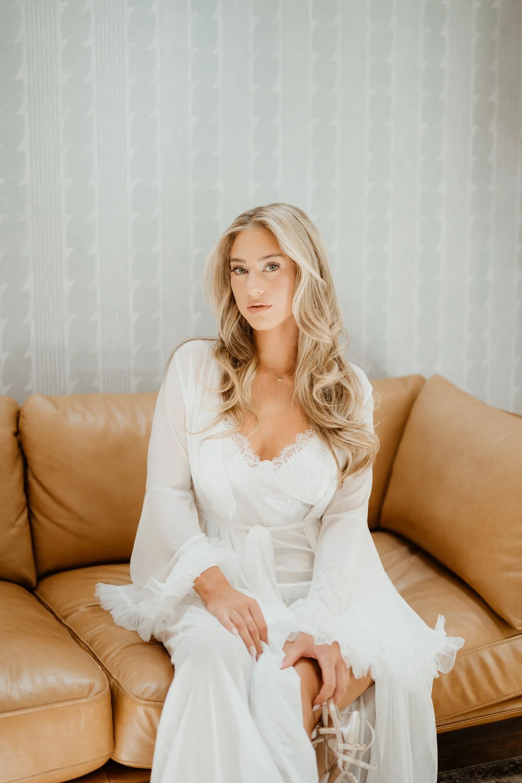 A woman in a white dress sitting on a tan leather couch, in a room with light-colored wallpaper.