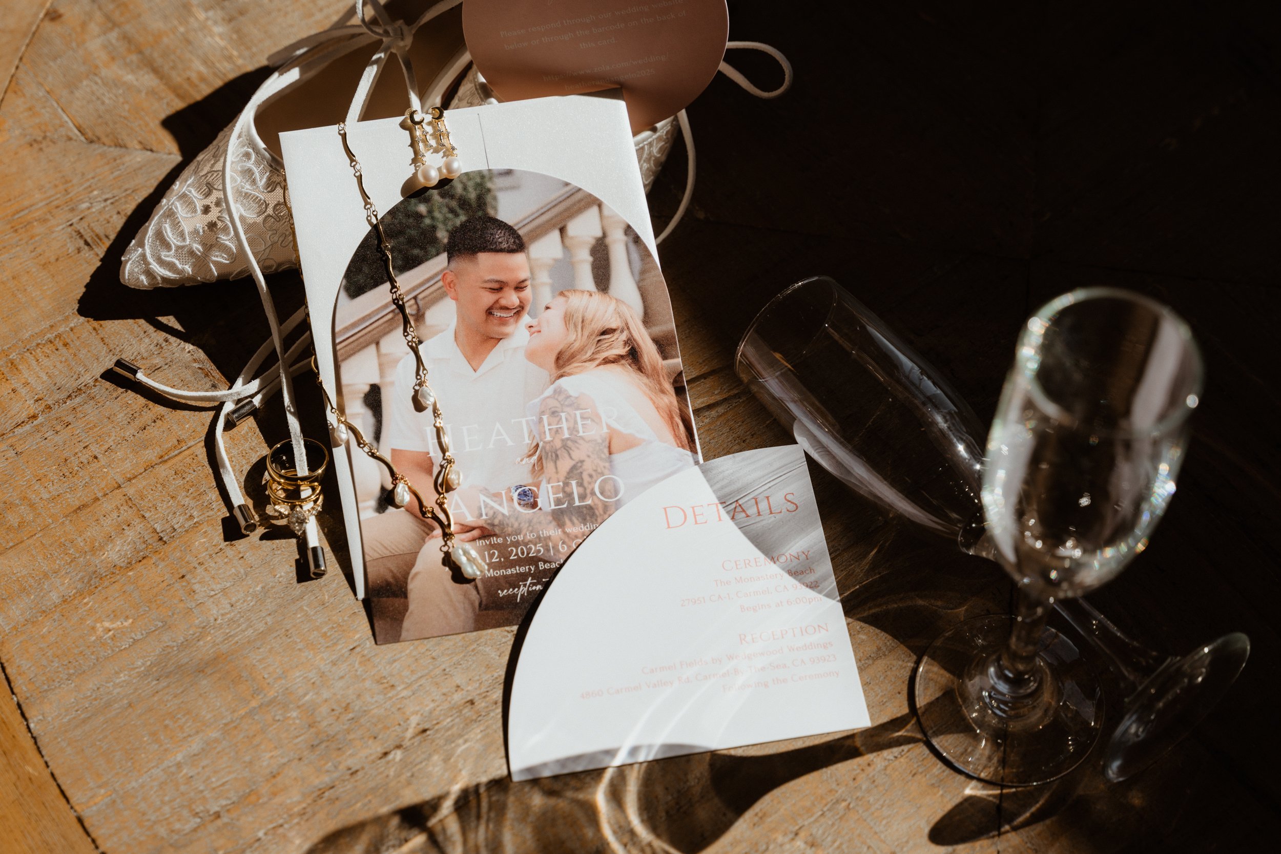 A wedding invitation card with a photo of a smiling couple, jewelry, and glasses of champagne on a wooden table. The invitation includes details about a wedding ceremony and reception.