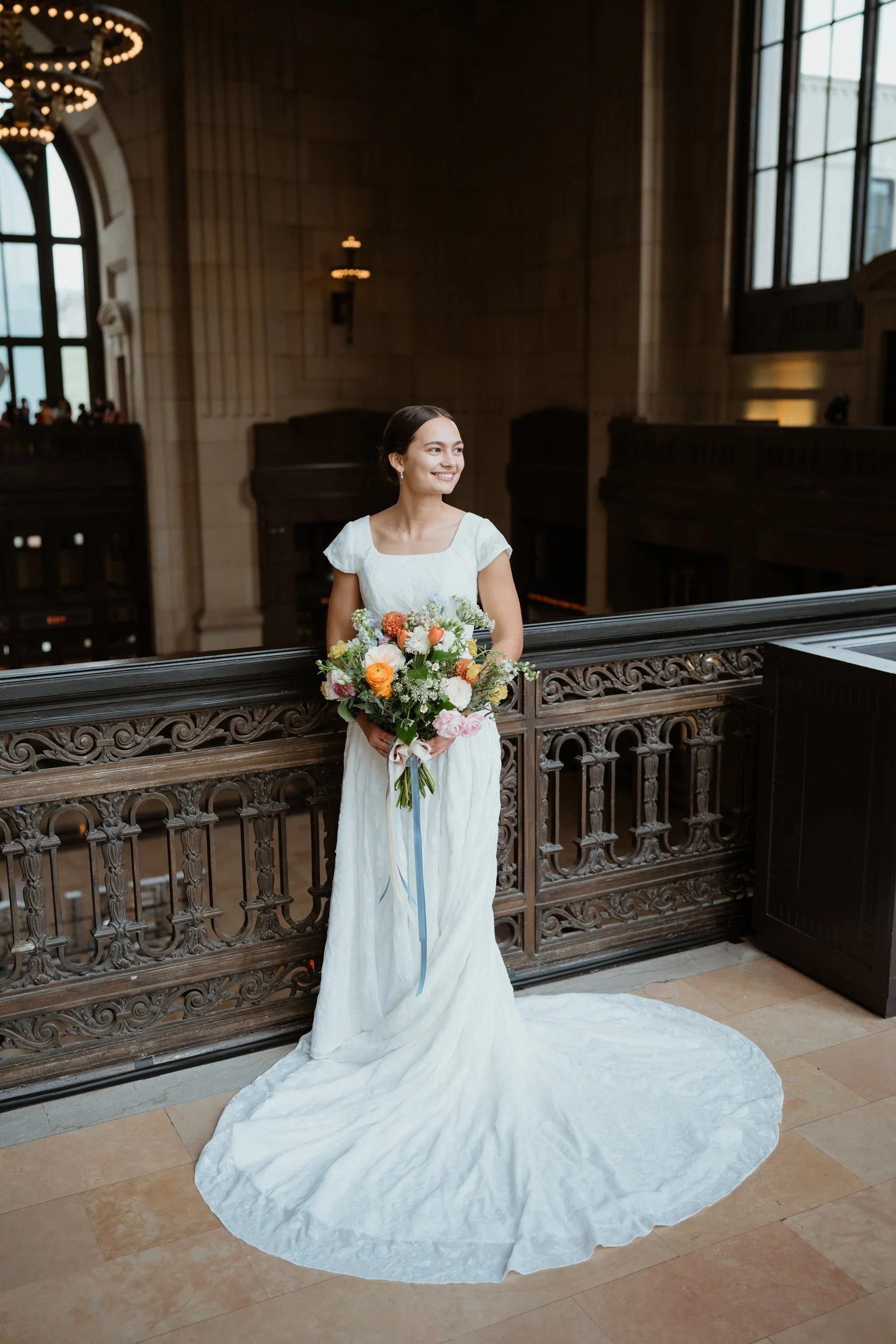 A bride in a white wedding dress holding a colorful bouquet of flowers, standing inside a large, ornate hall with high ceilings and large windows.