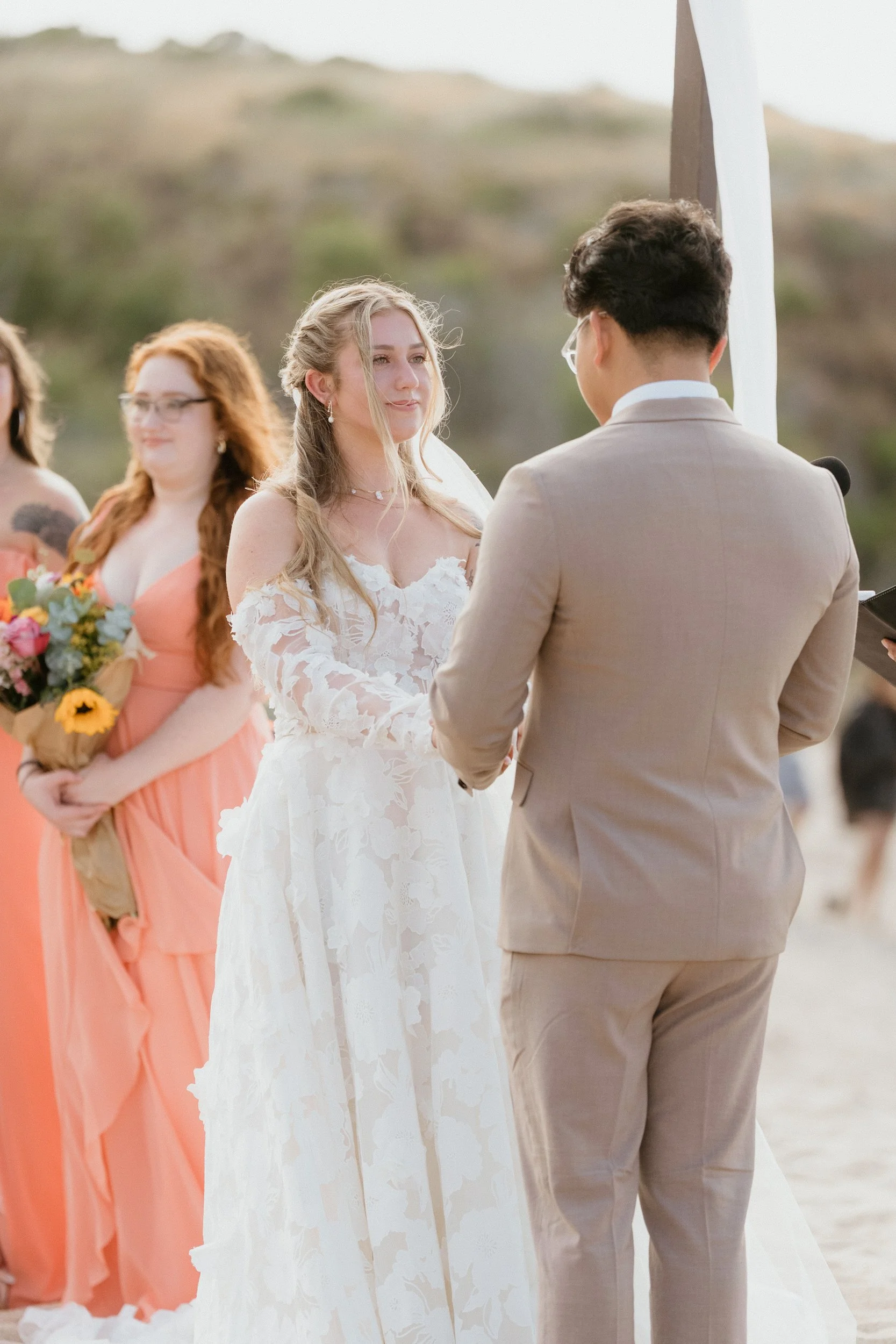 Bride and groom exchanging vows at outdoor beach wedding ceremony, with bridesmaids standing in the background holding flower bouquets.