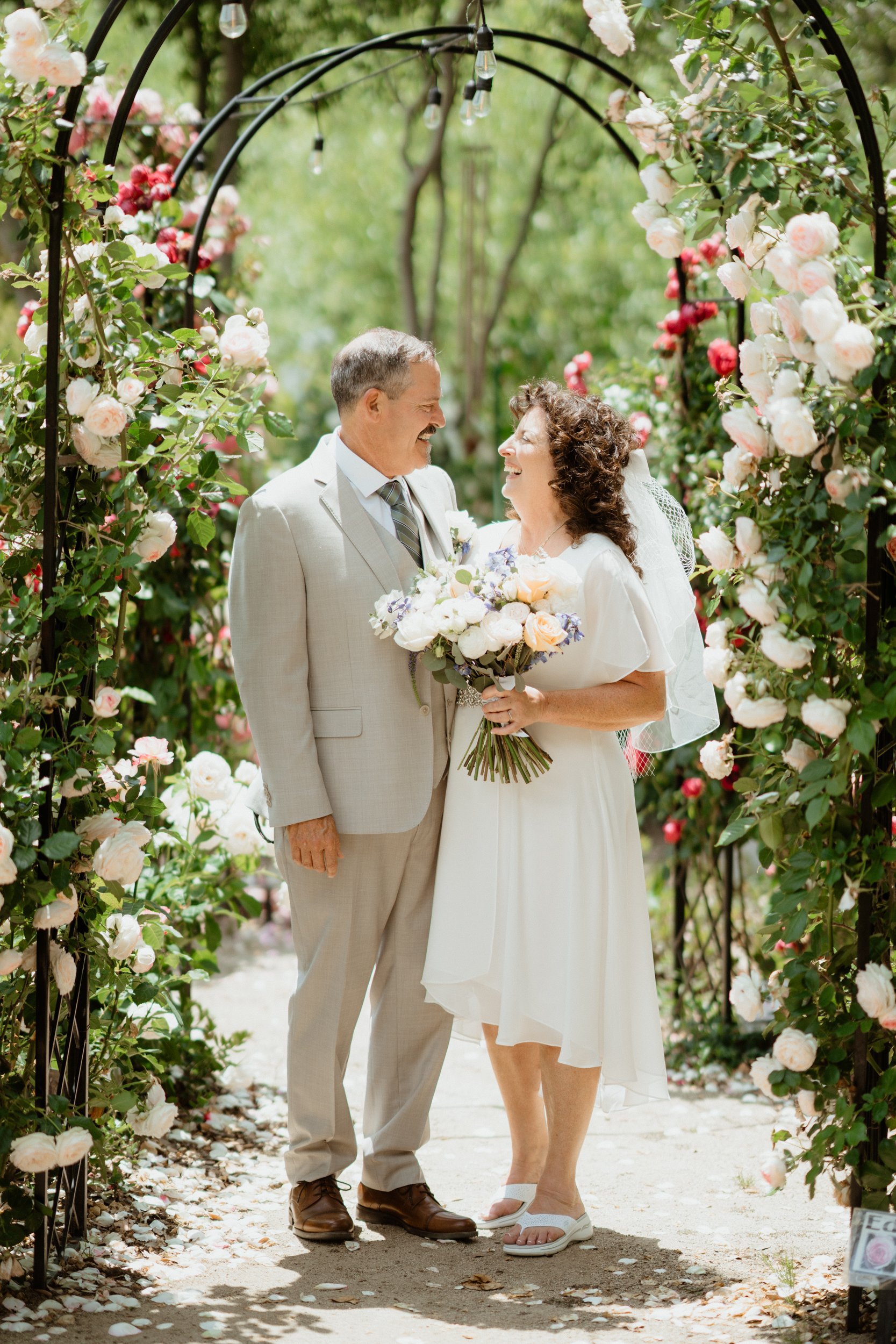 A bride and groom smiling at each other under a floral arch, with the bride holding a bouquet of flowers in a garden setting.