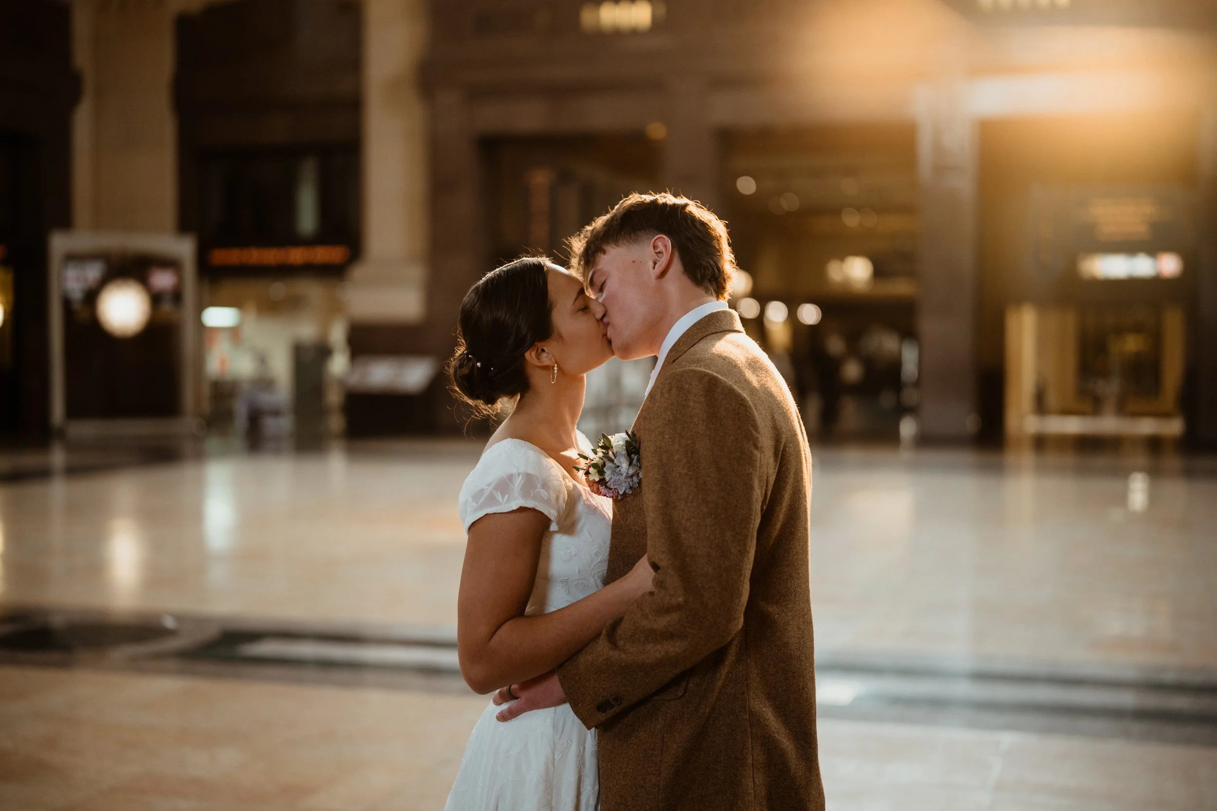 A couple kissing in a warmly lit indoor setting, with the woman wearing a white wedding dress and the man in a brown suit.