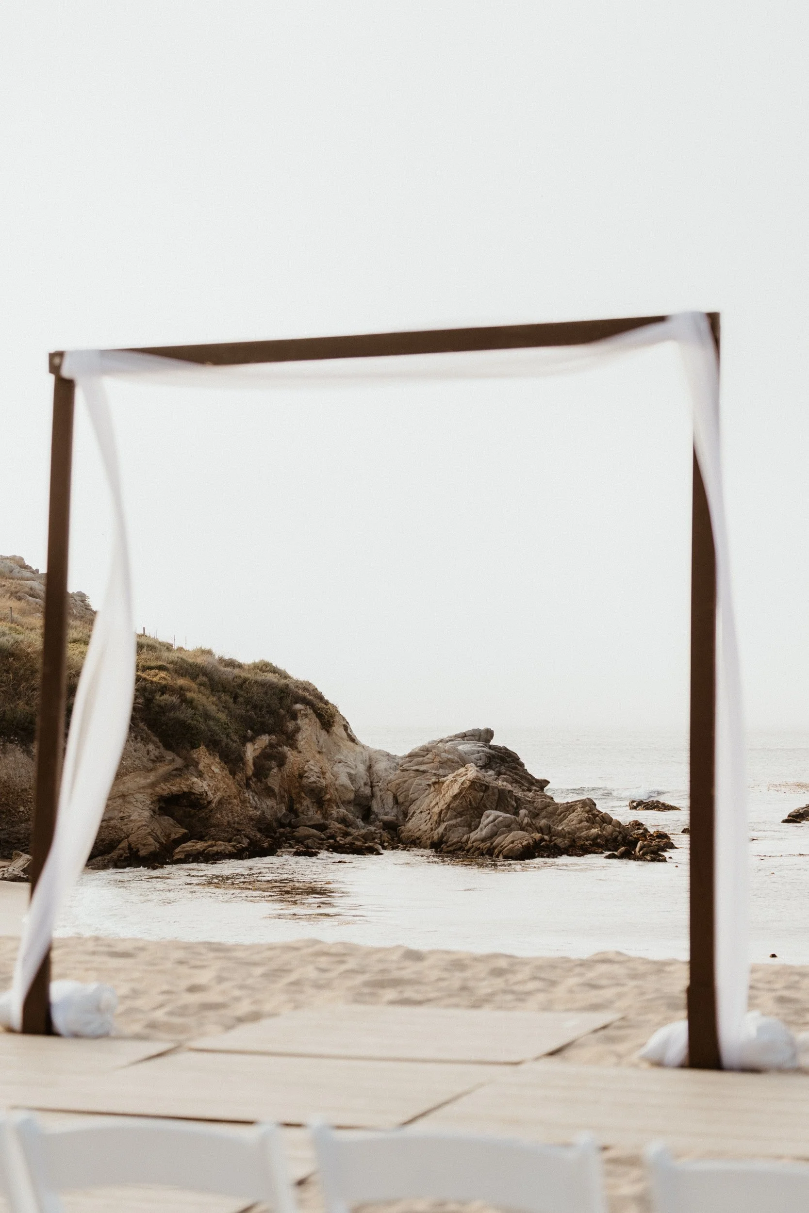 A wooden arch with white fabric draped from the top, set up on a sandy beach facing rocky cliffs and the ocean, under a foggy, overcast sky.