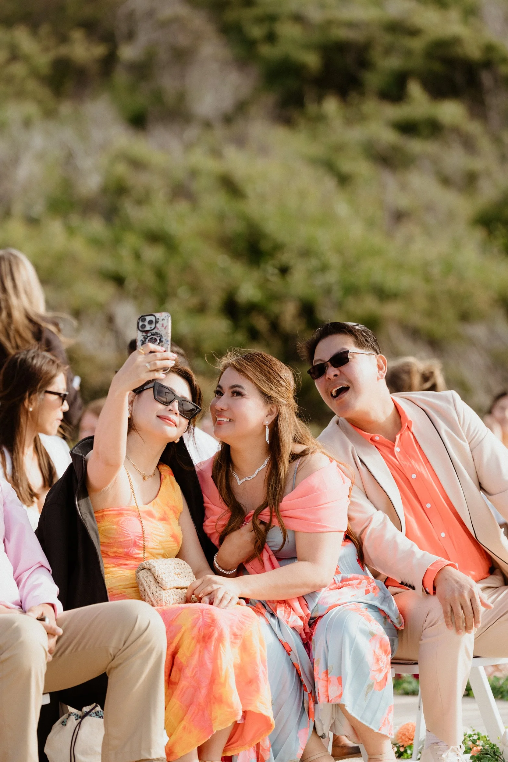 People taking a selfie at an outdoor event, sitting on chairs with a natural background.