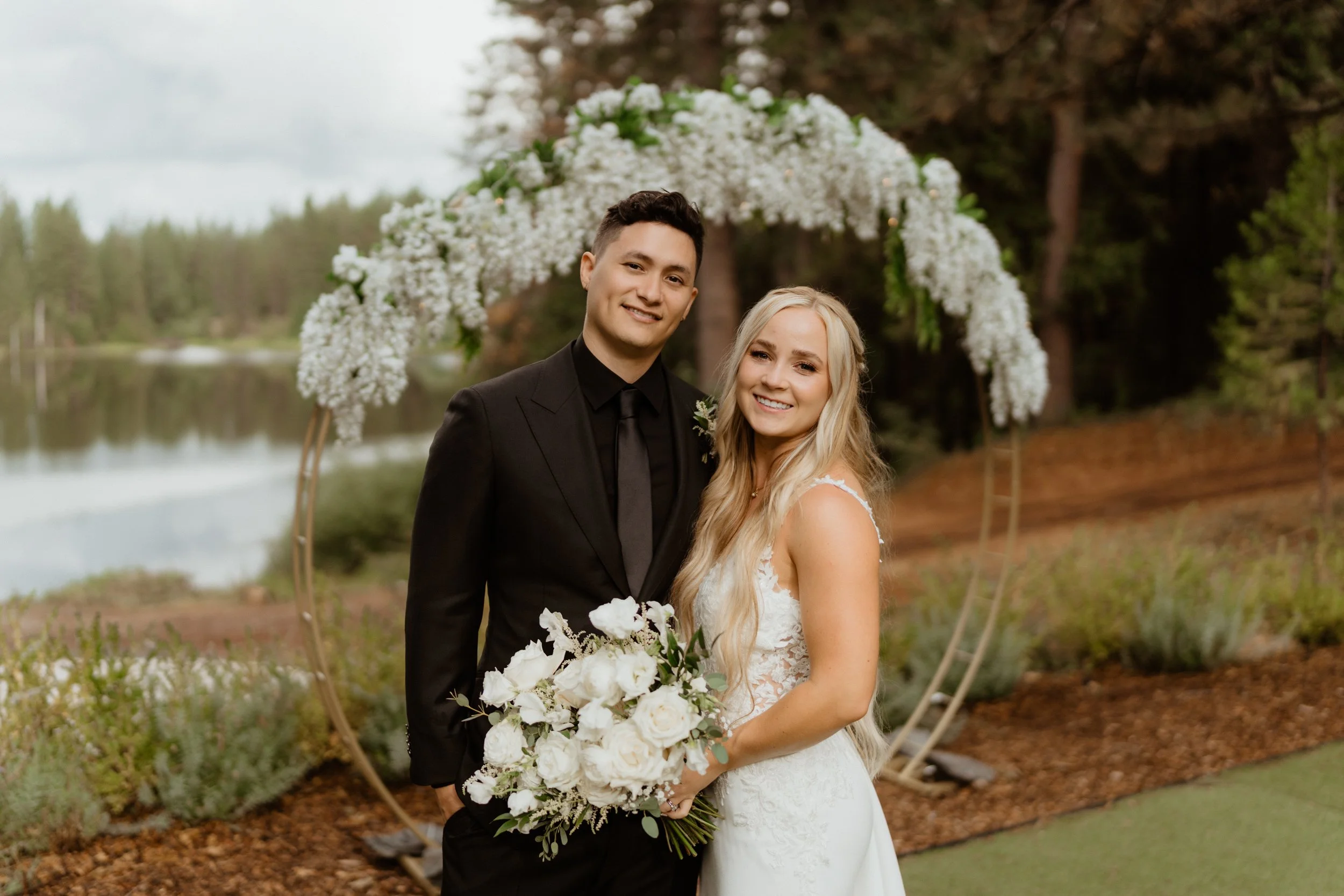 A bride and groom standing together outdoors during their wedding, in front of a white floral arch near a lake and forest.