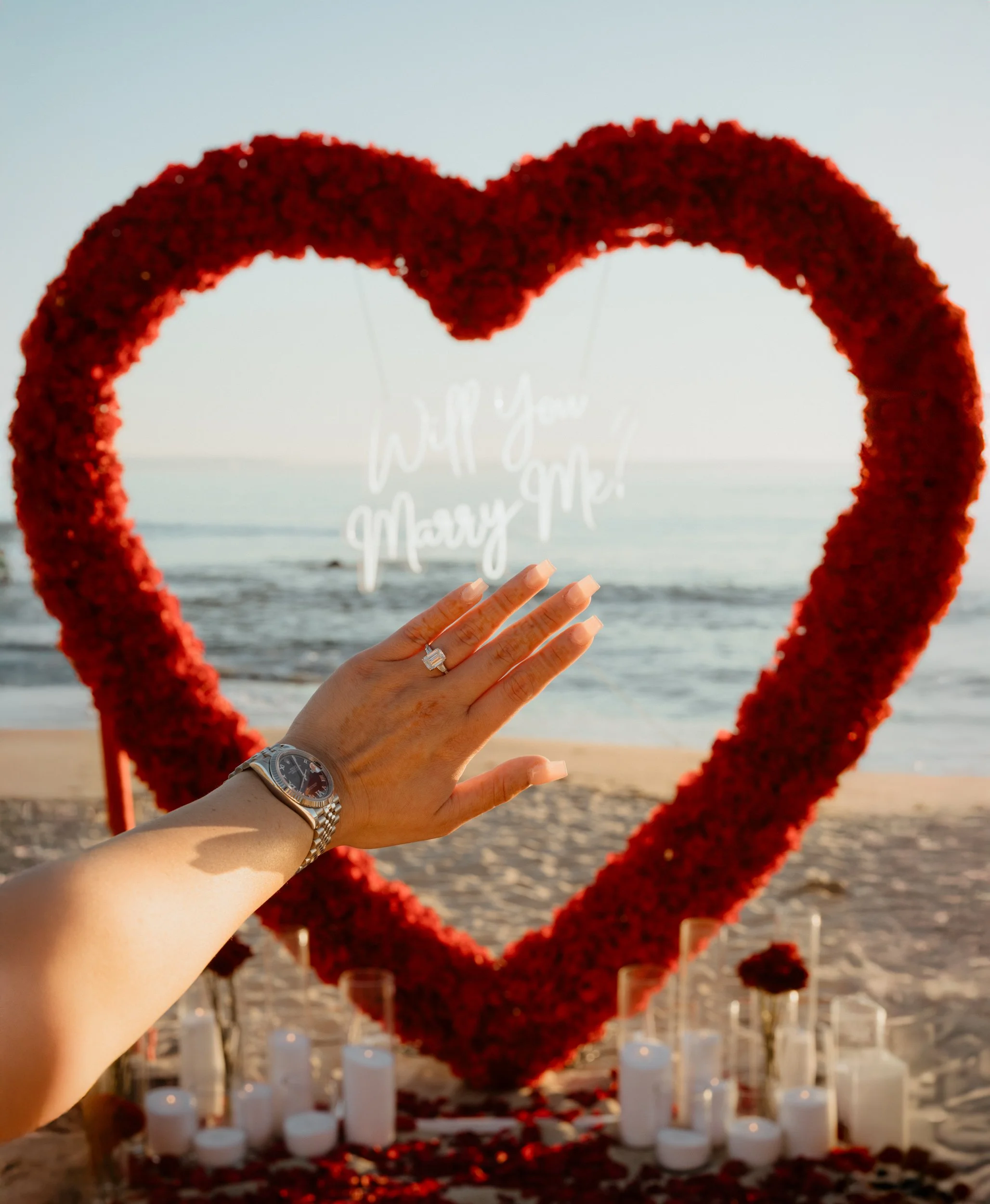 A hand with a wedding ring and watch reaching out towards a heart-shaped floral wreath with candles and rose petals at the beach during sunset, with text 'Will you marry me?' in the background.