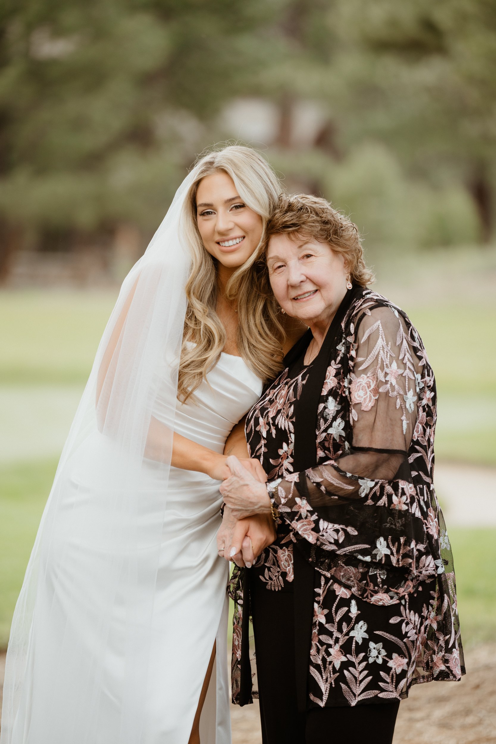 A bride in a white wedding dress and veil holding hands with an elderly woman in a black floral blouse, both smiling outdoors in a park.