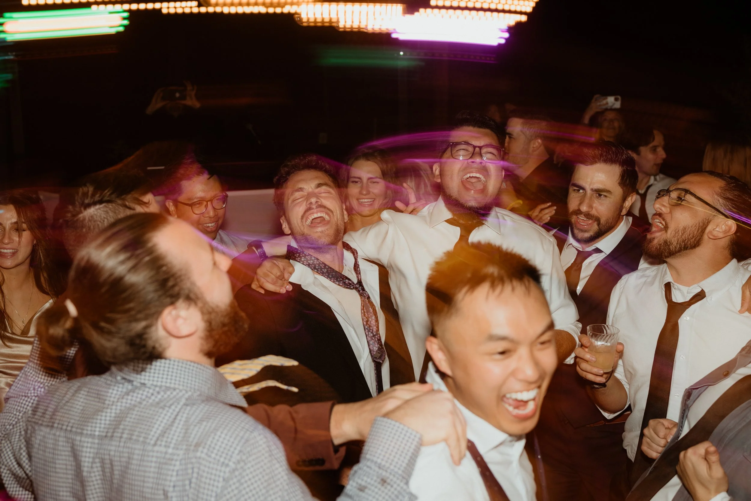 Group of diverse friends at a celebration, laughing, dancing, and enjoying each other's company, with colorful blurred lights in the background.
