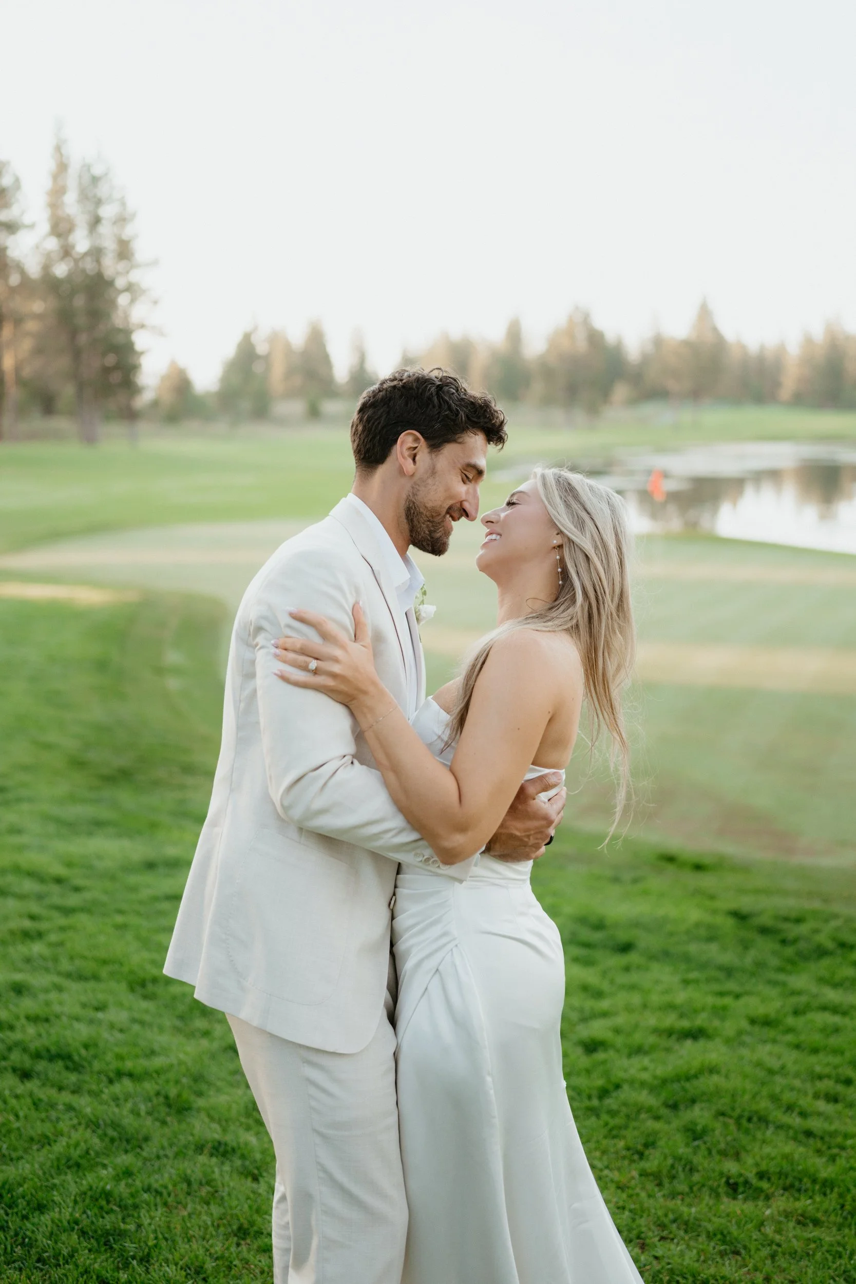 A happy couple in wedding attire embracing outdoors on a grassy field with trees and a lake in the background.