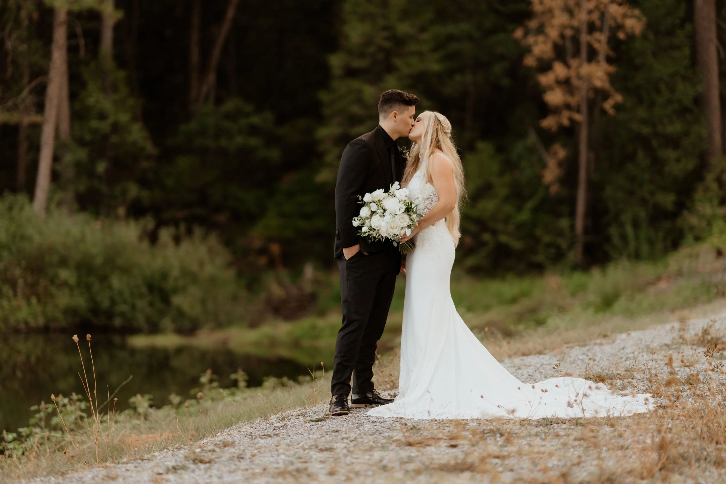 A bride and groom kissing outdoors near a river, with the bride holding a bouquet of white flowers and trees in the background.
