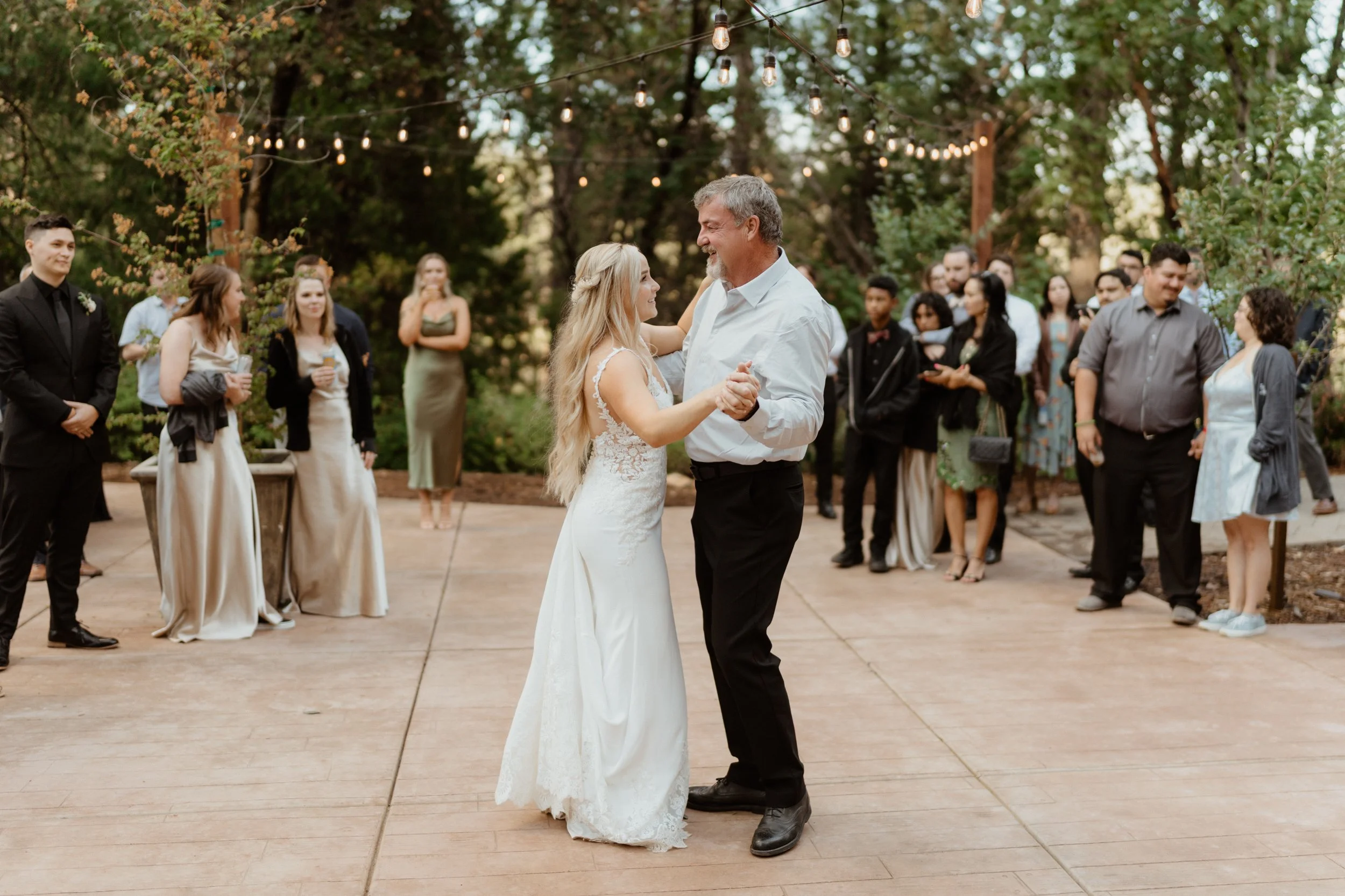 A bride and a man dancing together on a wooden dance floor at an outdoor wedding reception with guests around, string lights hanging in a wooded area.