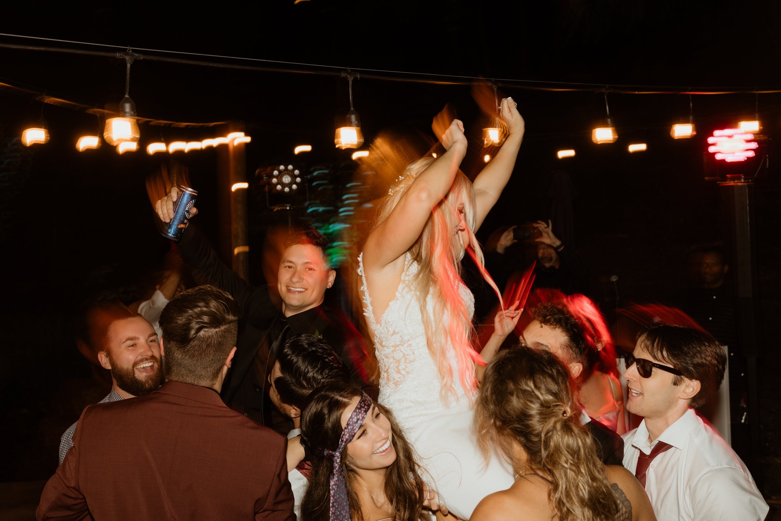 People dancing and celebrating at a party, with a woman in a white dress lifted into the air while others cheer around her, in a dimly lit venue with string lights.