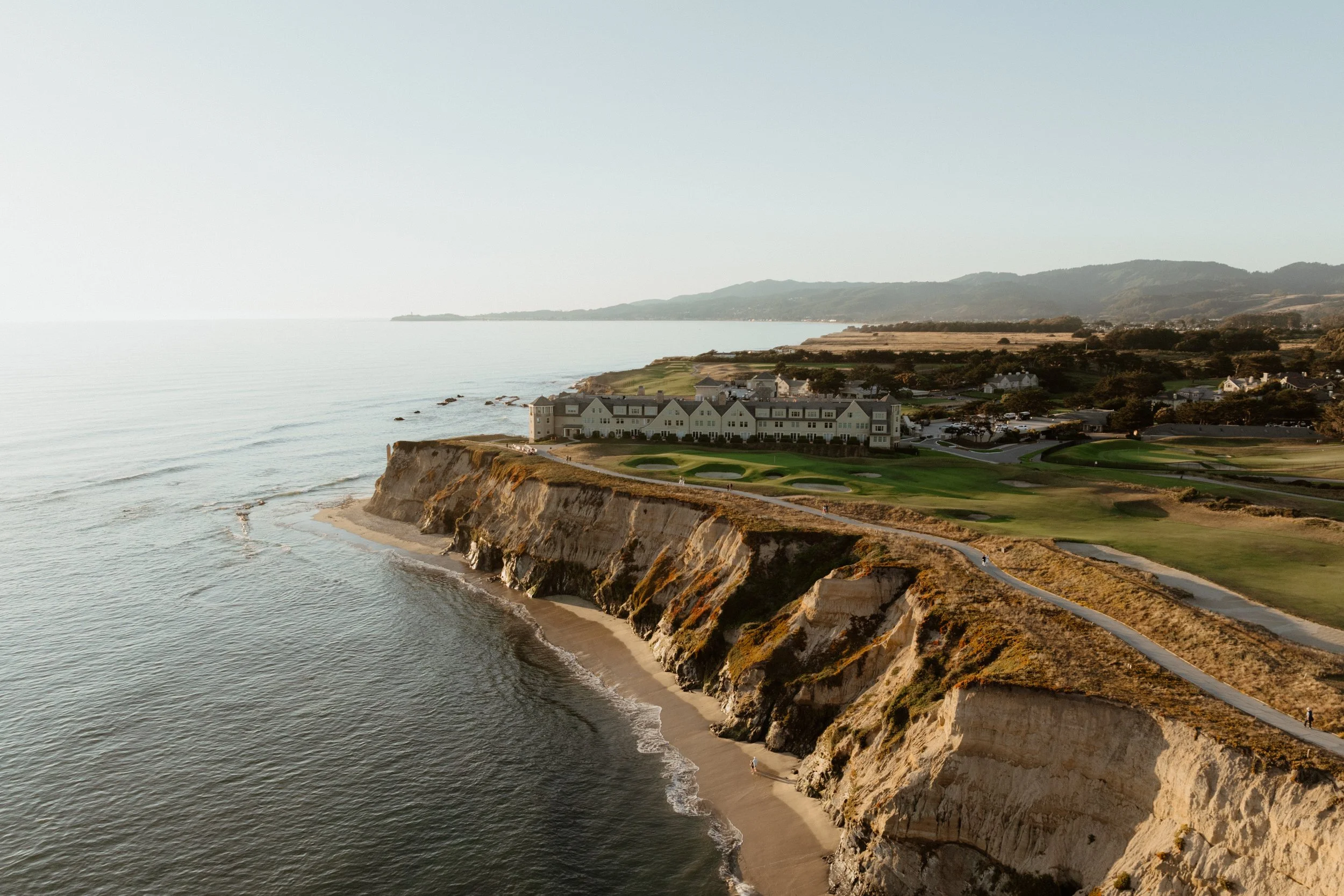 Coastal cliffs overlooking a golf course with a large white building by the shoreline at sunset.
