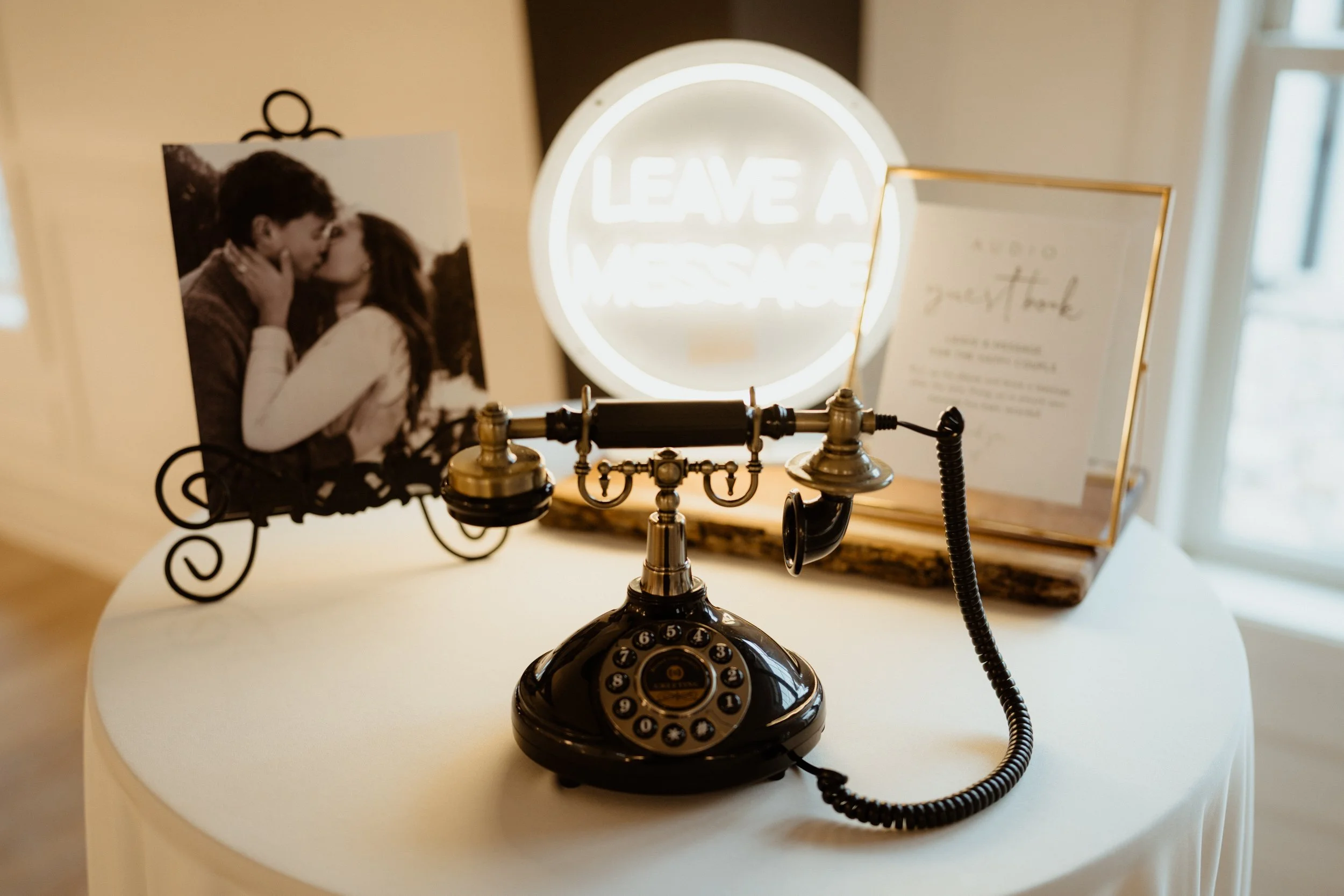 Black vintage rotary phone on white table with wedding photo, neon sign that says 'Leave a Message', and framed wedding message signs in background.
