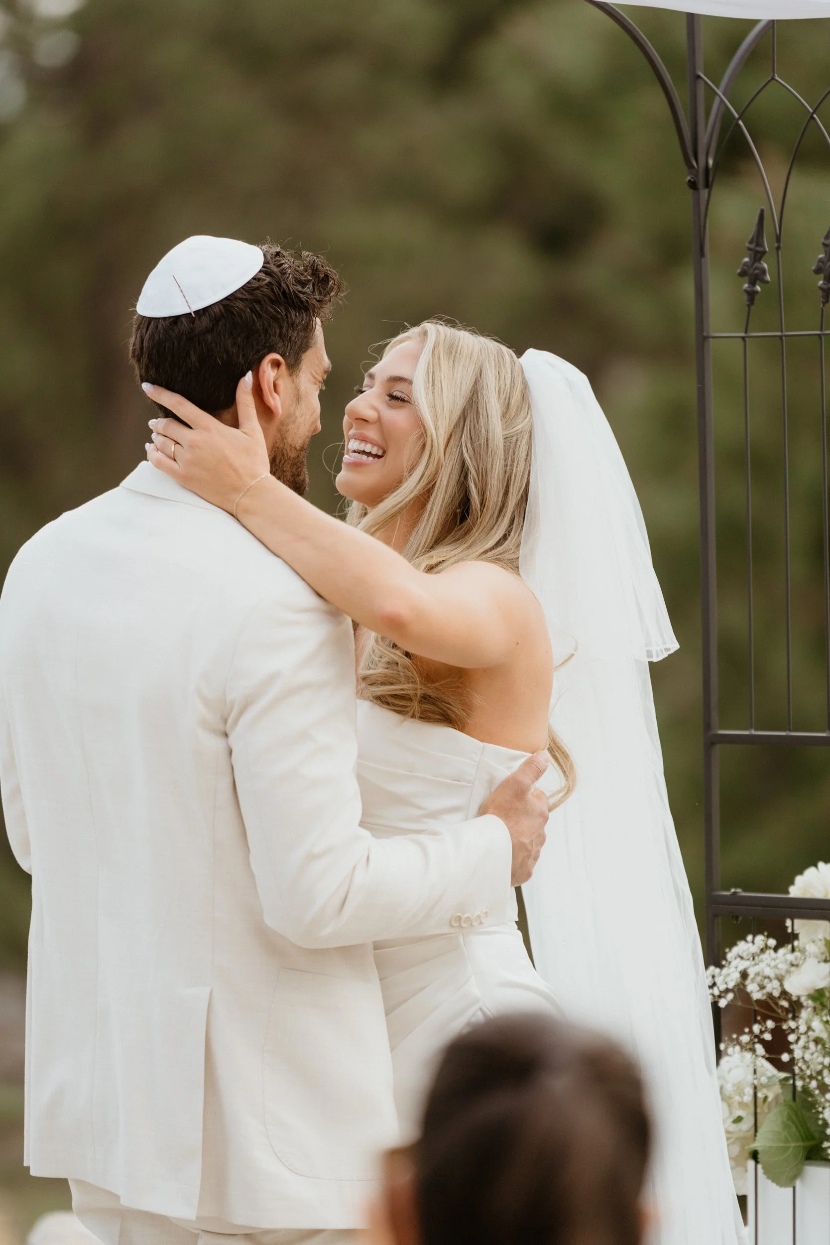 A bride and groom are embracing outdoors during their wedding ceremony, with the bride smiling joyfully and wearing a veil, and the groom wearing a yarmulke and white suit.