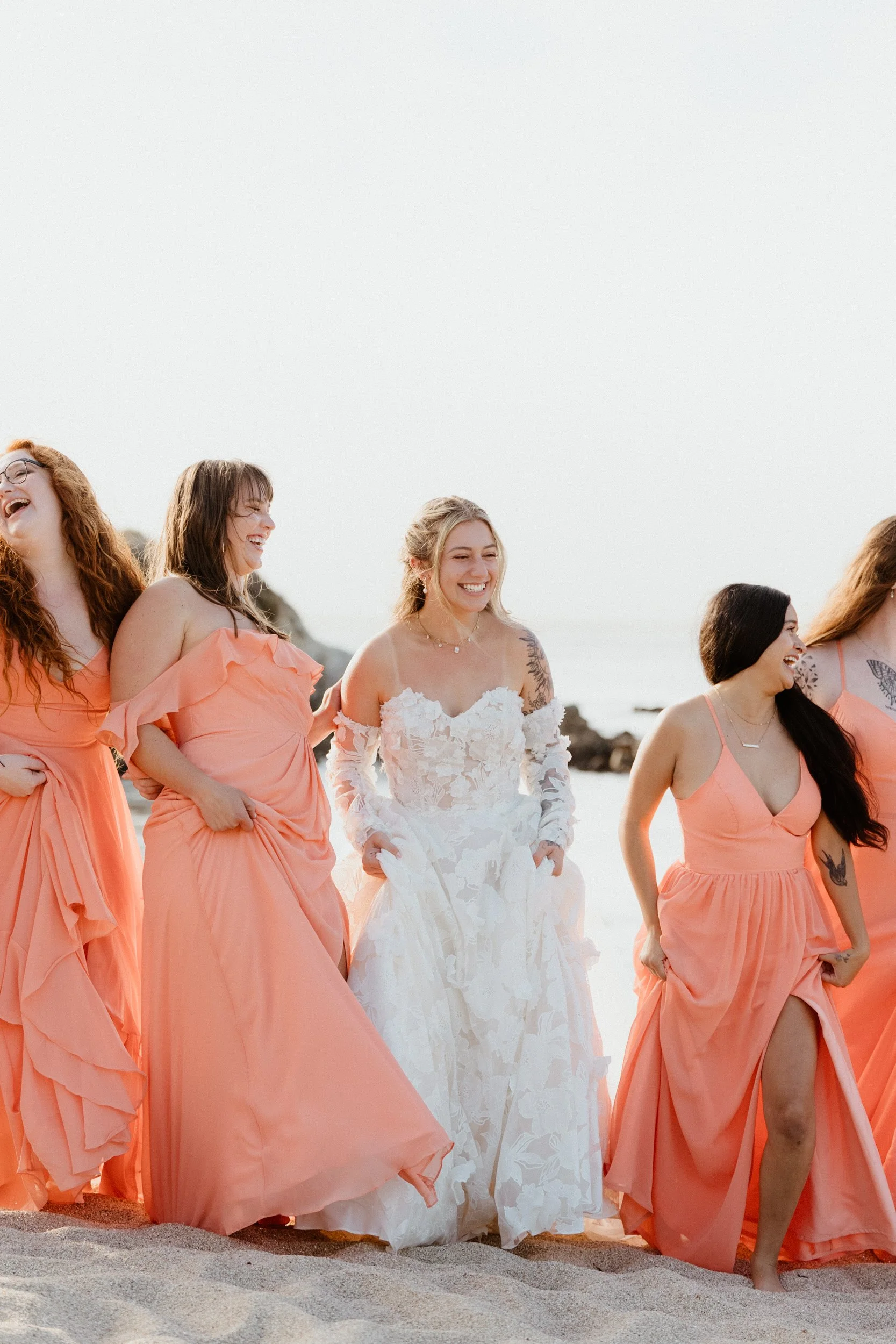 Group of women in peach and white dresses, laughing and walking on the beach at sunset.