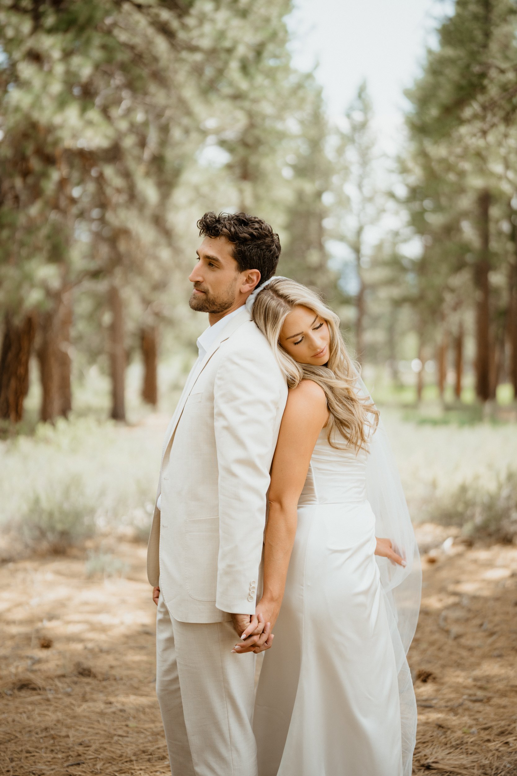 A man and woman in wedding attire standing back-to-back in a forest, holding hands, with the woman resting her head on the man's shoulder.